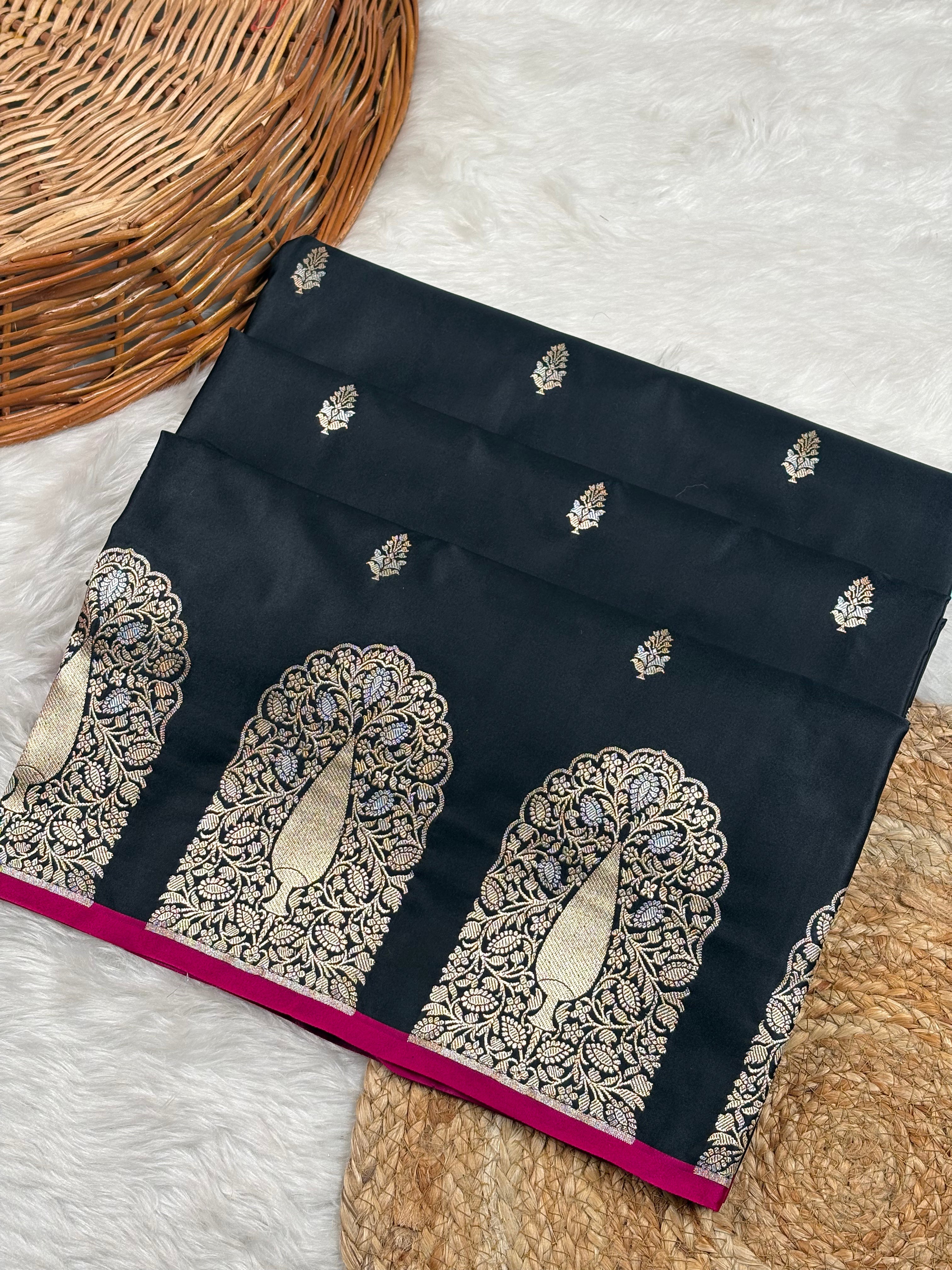 A folded black mashru katan saree with ornate alfy mina and floral border weaving, displayed on a white faux fur surface with a wicker basket and woven mat as props.