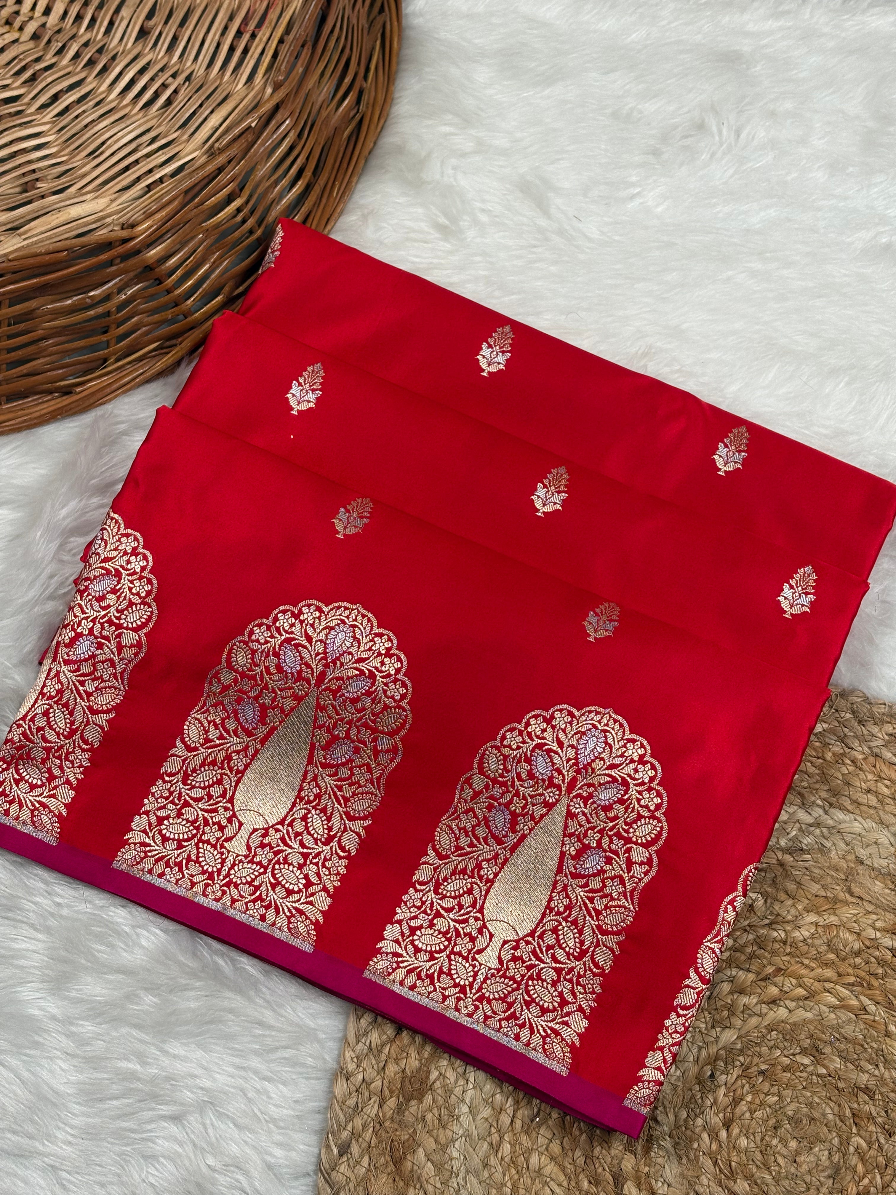 A folded red silk saree with intricate sona rupa zari patterns, displayed on a white faux fur surface next to a woven wicker basket.