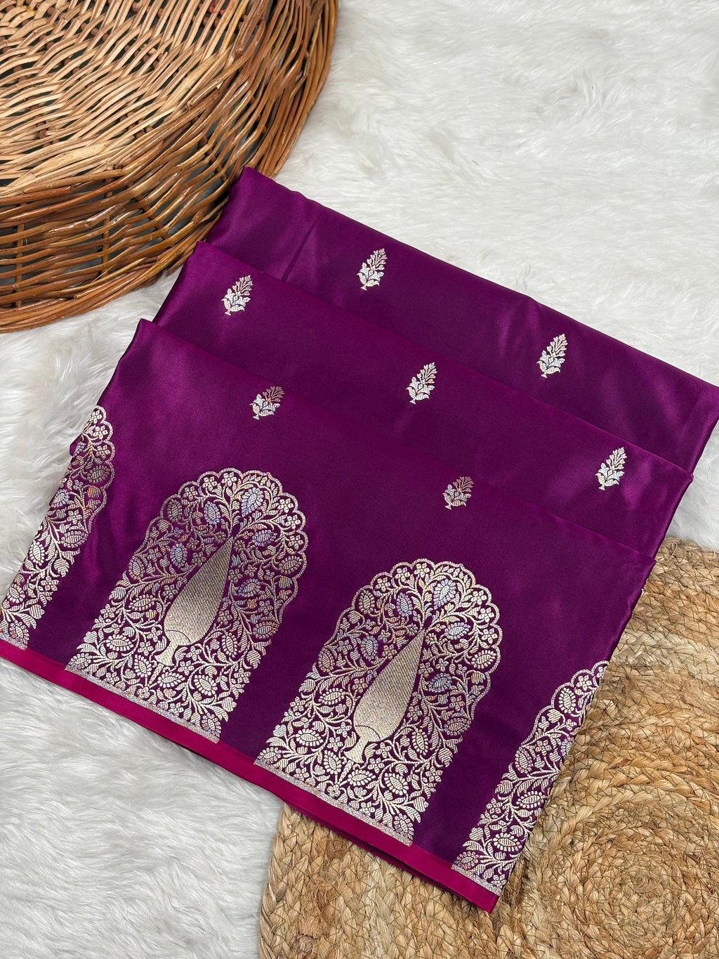 A folded deep purple silk saree with intricate alfi mina weaving, featuring a border with mehrab motifs, styled on a white faux fur surface with a woven basket.