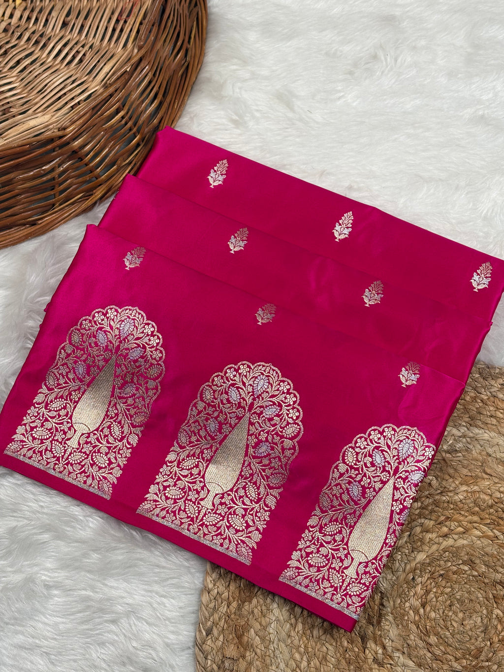 A folded magenta pink saree with alfi mina mehrab border and floral motifs weaving, displayed on a white faux fur surface with a woven basket and jute mat as props.