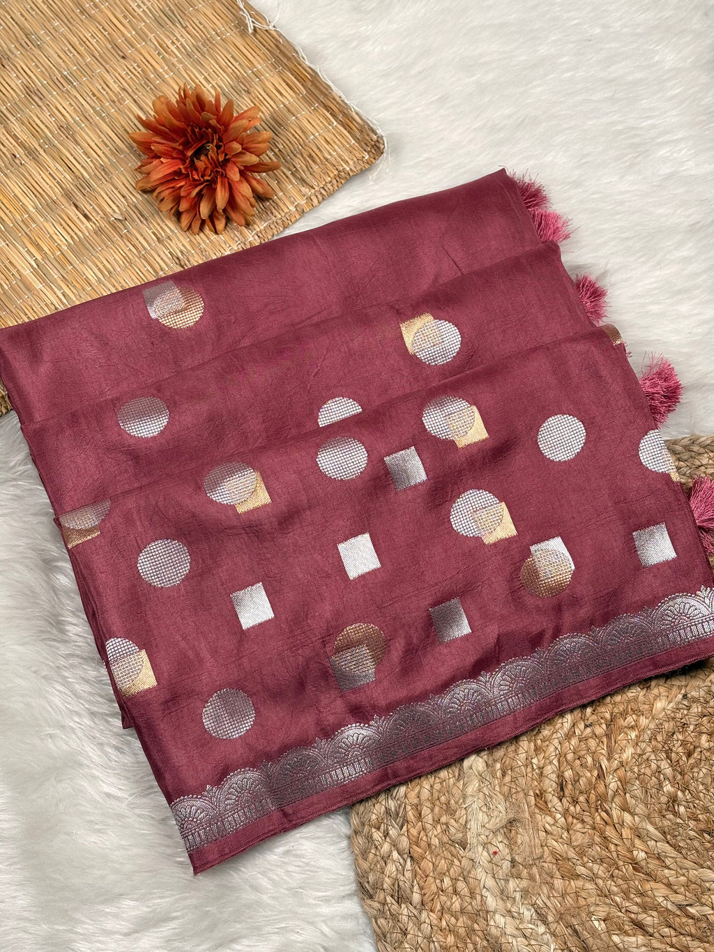 A folded maroon chinya silk saree with alfi mina geometric motifs and a scalloped border weaving, styled on a white faux fur surface with a woven mat and a dried orange flower.