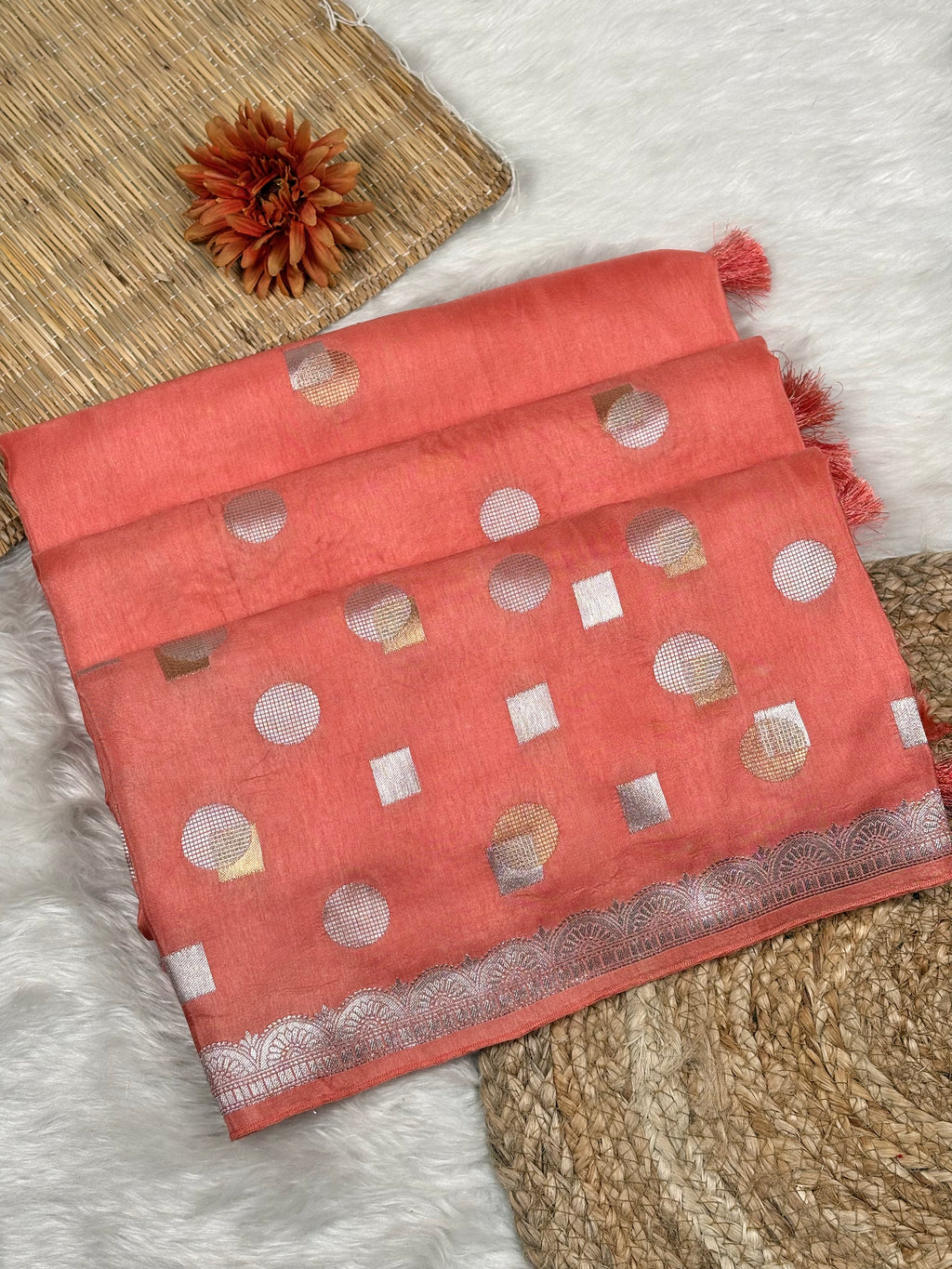 A coral pink saree with alfi mina geometric motifs and a zari border, styled on a white faux fur rug with a woven mat and a dried flower.