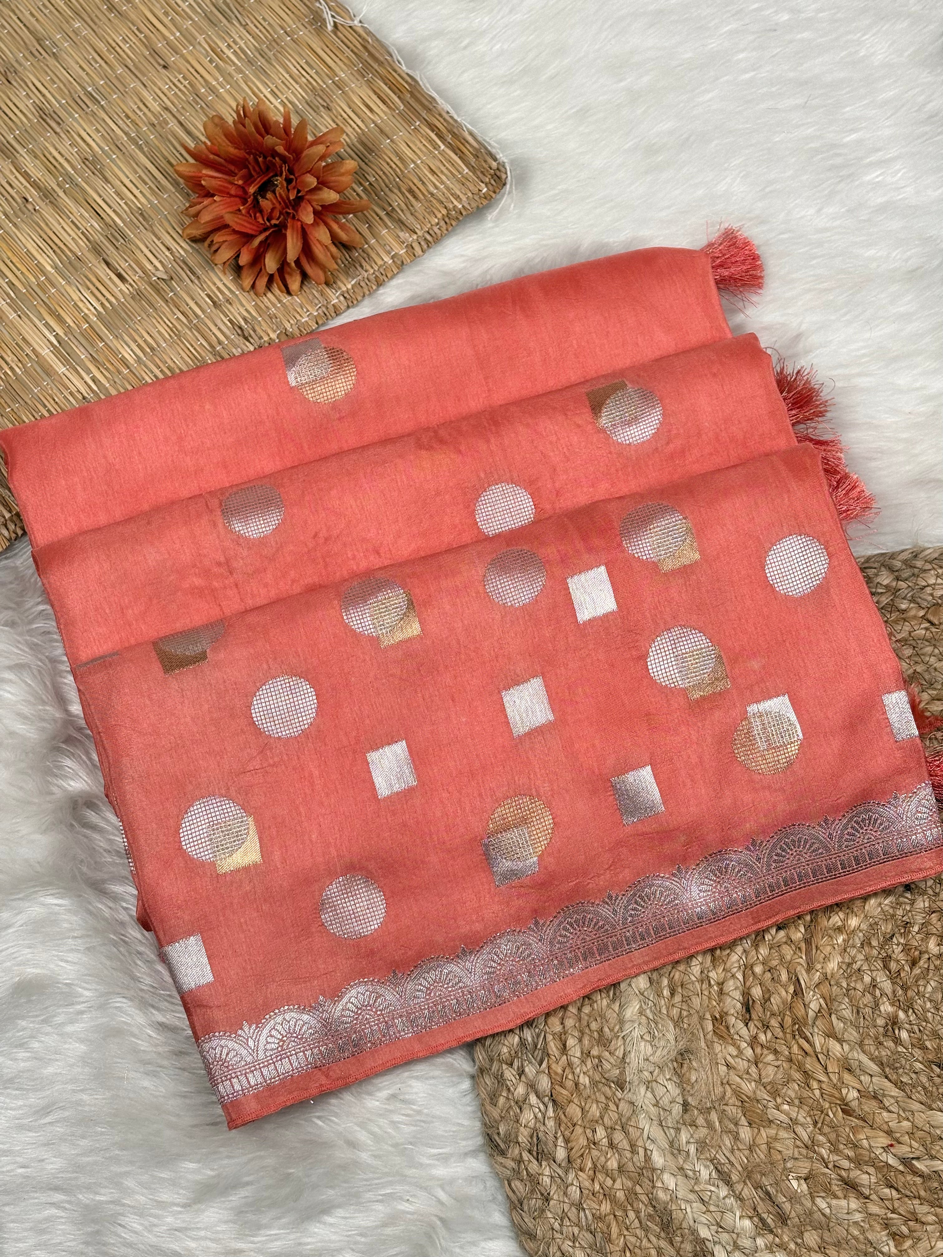 A coral pink saree with alfi mina geometric motifs and a zari border, styled on a white faux fur rug with a woven mat and a dried flower.