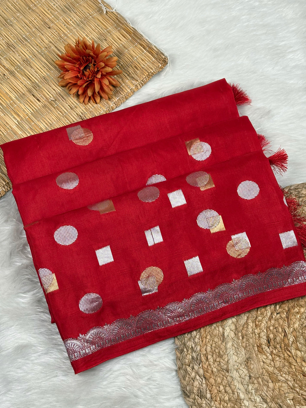 A folded red saree piece with alfi mina woven motifs, displayed on a white faux-fur surface with woven mat props and a dried orange flower.