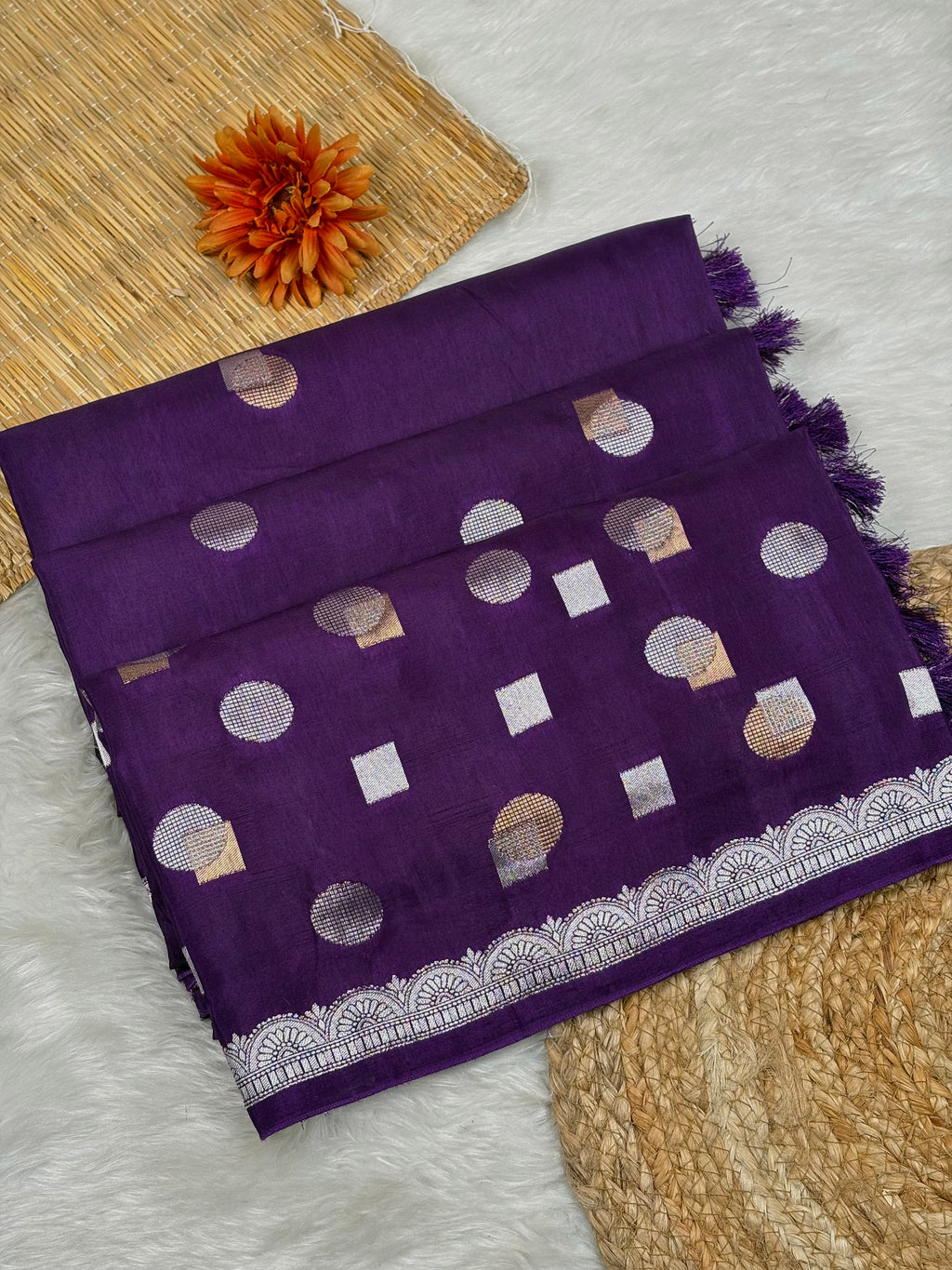 A folded purple chinya silk saree with alfi mina geometric motifs and a silver border weaving, displayed on a white faux fur surface with decorative woven mats and an orange flower.