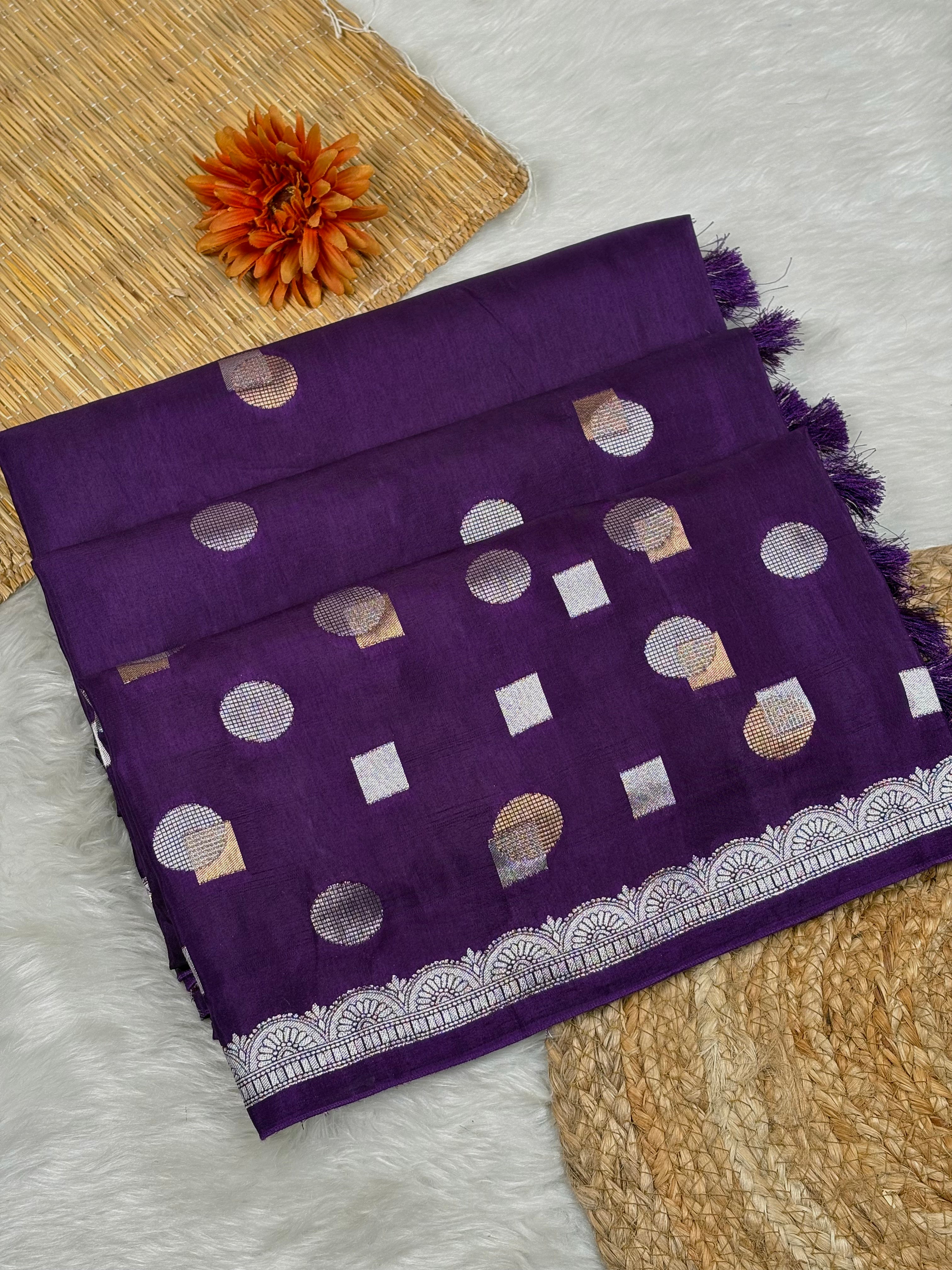 A folded purple chinya silk saree with alfi mina geometric motifs and a silver border weaving, displayed on a white faux fur surface with decorative woven mats and an orange flower.