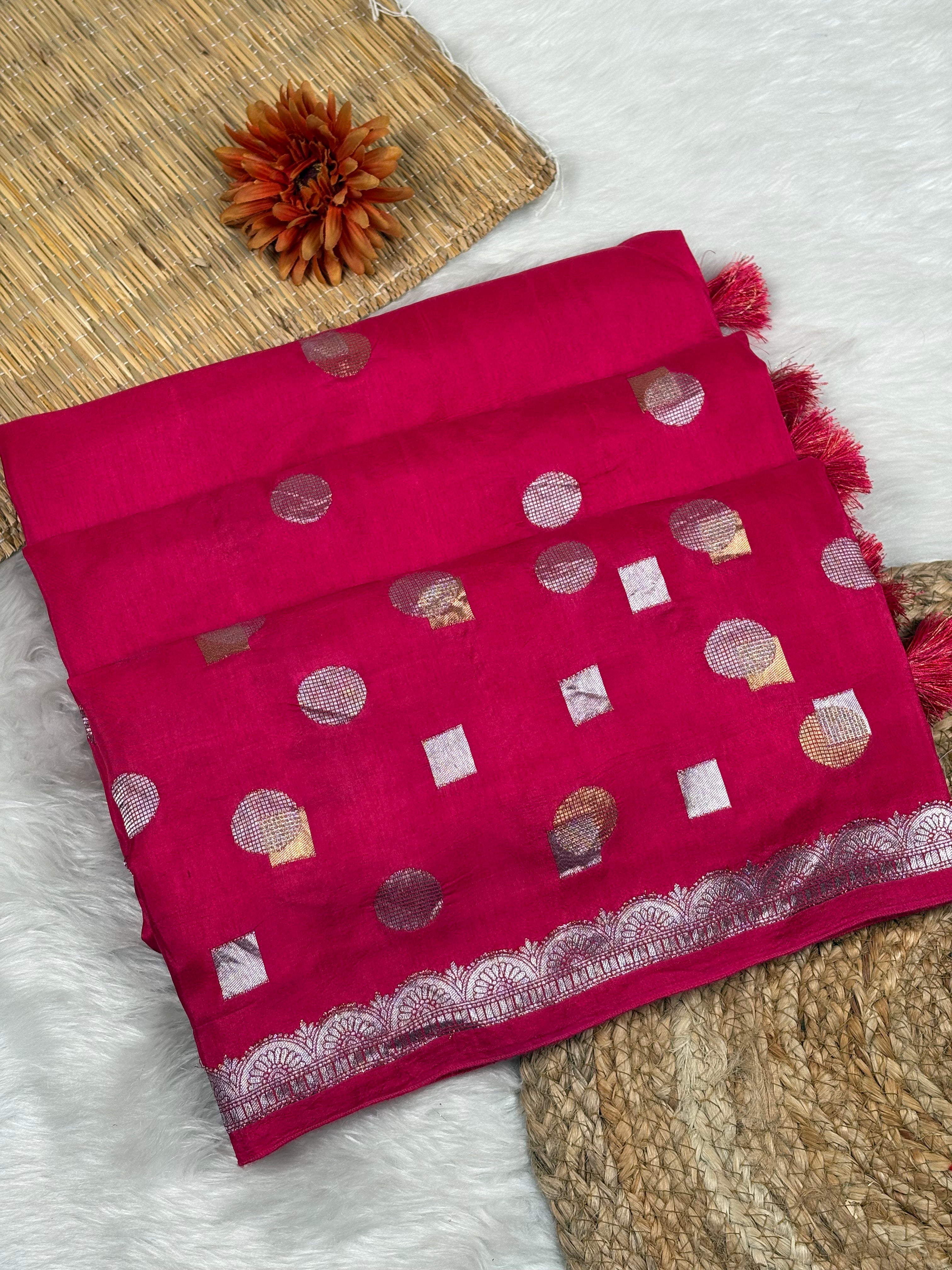 A folded deep pink saree with alfi mina woven motifs, displayed on a white faux fur surface with a woven mat and a dried orange flower as props.