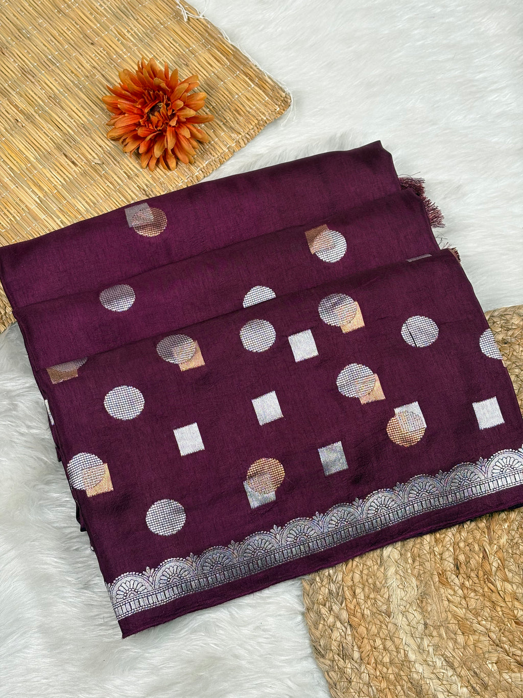 A folded deep purple saree with alfi mina woven motifs, displayed on a white faux fur surface with a woven mat and orange flower prop.