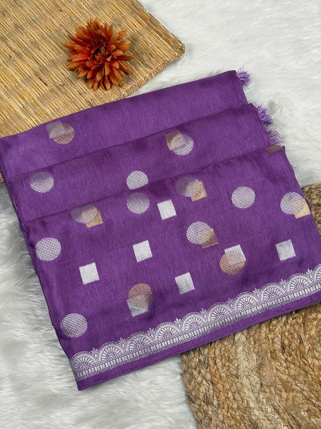 A folded purple chinya silk saree, featuring alfi mina geometric motifs and a zari weaving border, displayed on a white faux fur surface with a woven mat and a dried orange flower.
