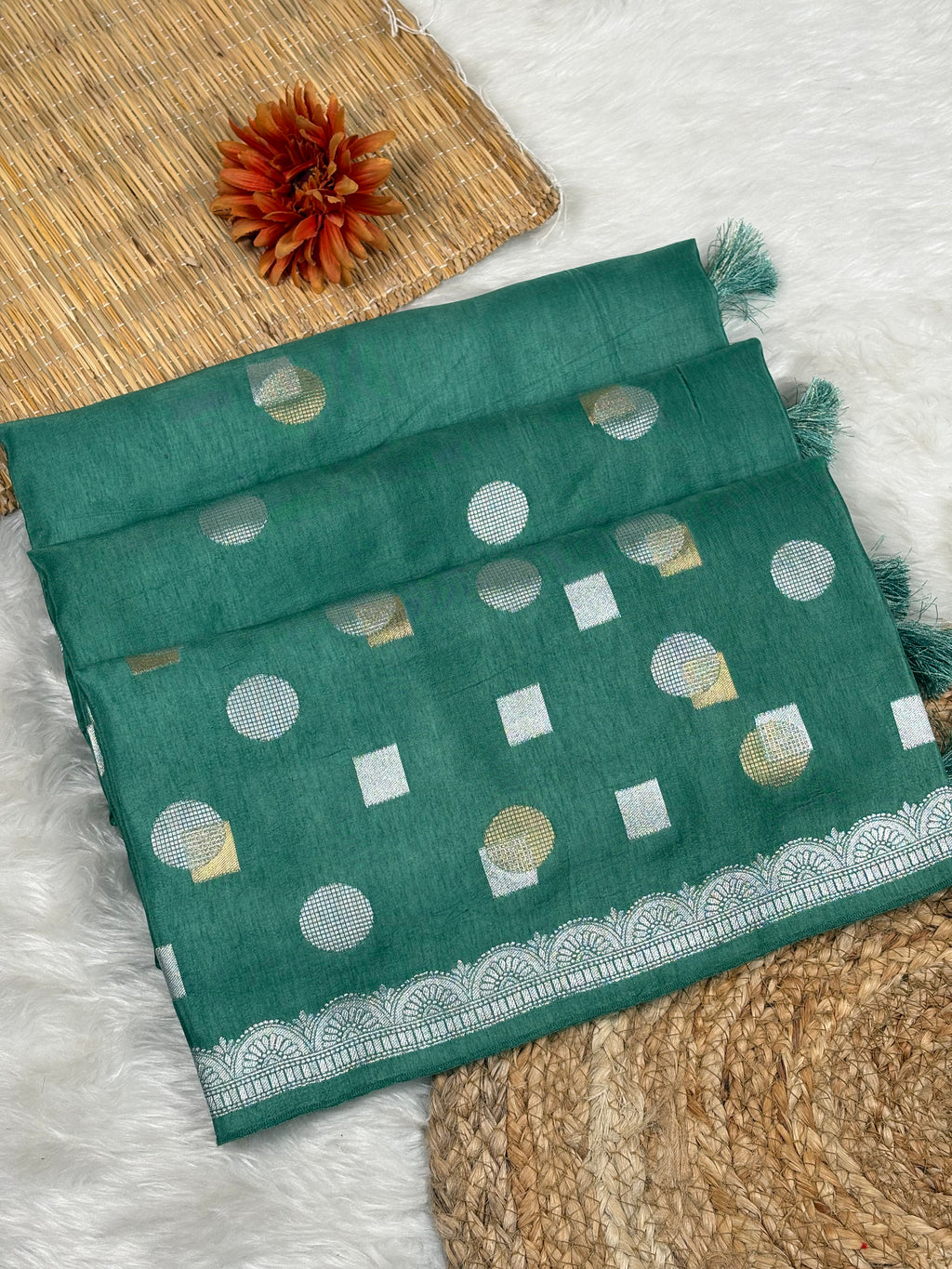 A folded dark teal green saree with alfi mina geometric motifs and a zari weaving border, styled on a white faux-fur surface with a woven mat and dried orange flower.