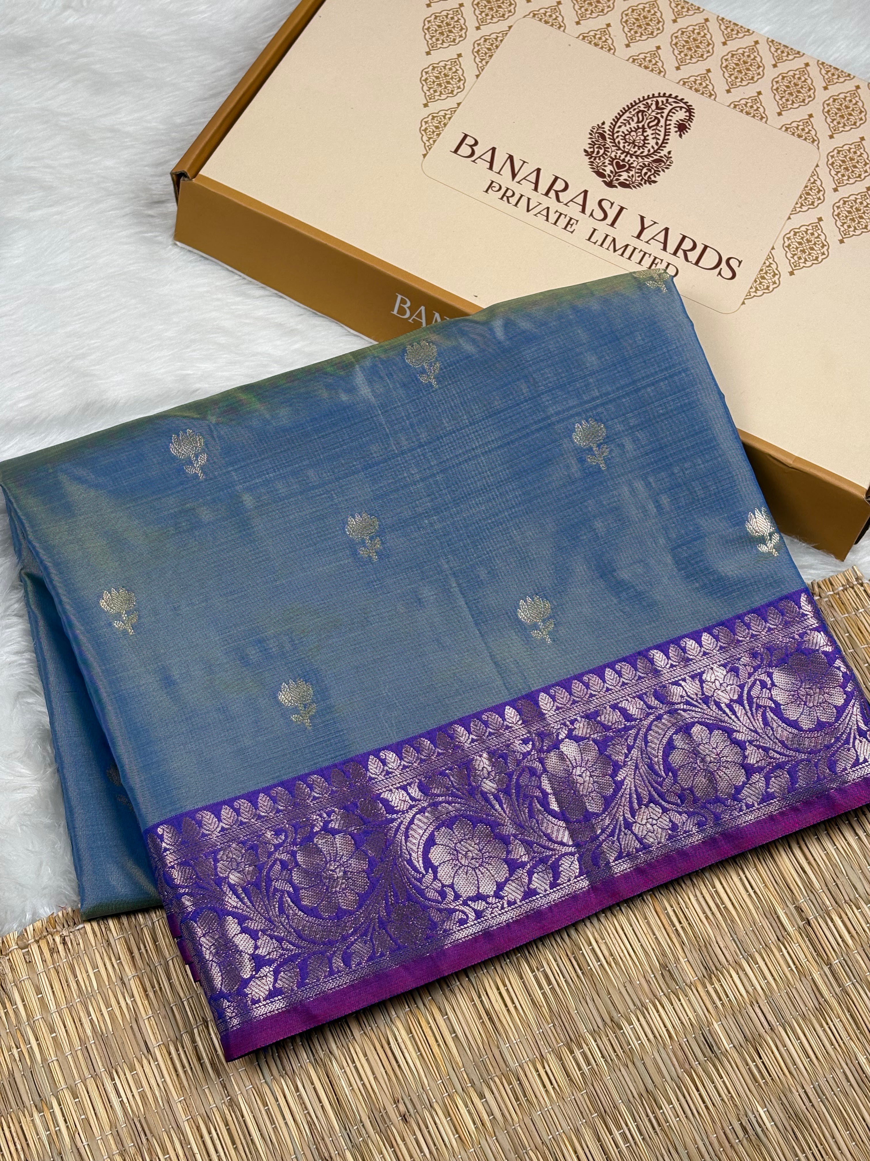 A folded blue saree with a purple border and gold floral motifs, displayed next to its beige packaging box labeled 'Banarasi Yards Private Limited' on a woven mat.