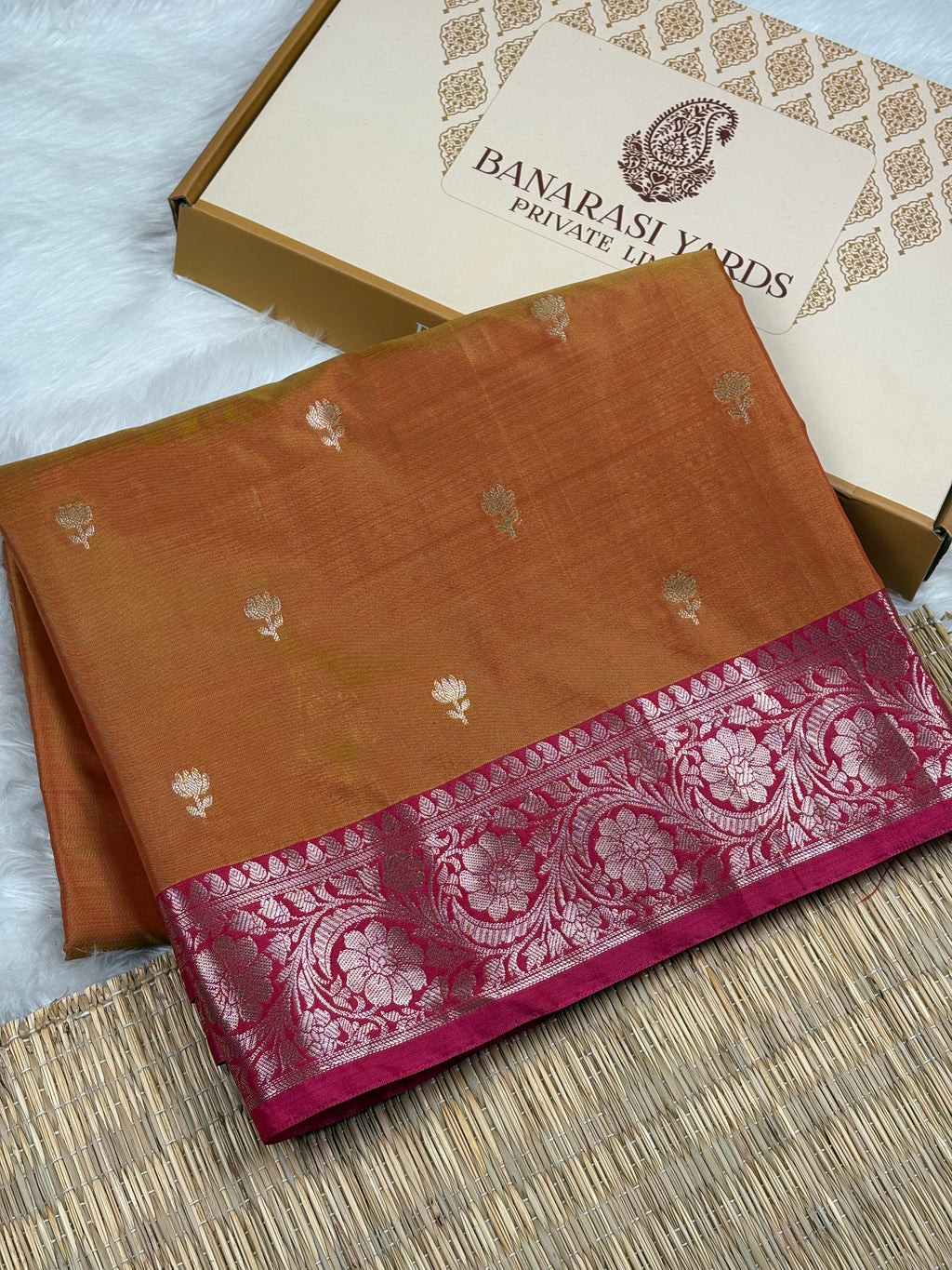 A folded Banarasi silk saree in a warm orange-brown shade with small zari floral motifs and a wide magenta-pink border featuring intricate floral weaving, displayed next to its branded packaging box.