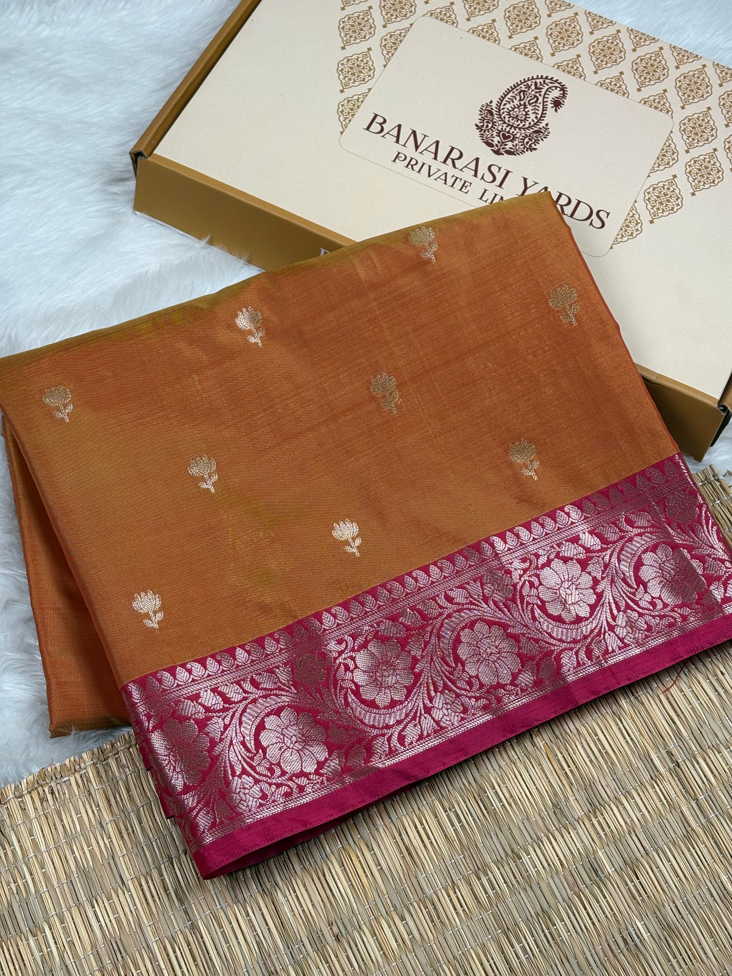 A folded Banarasi silk saree in a warm orange-brown shade with small zari floral motifs and a wide magenta-pink border featuring intricate floral weaving, displayed next to its branded packaging box.