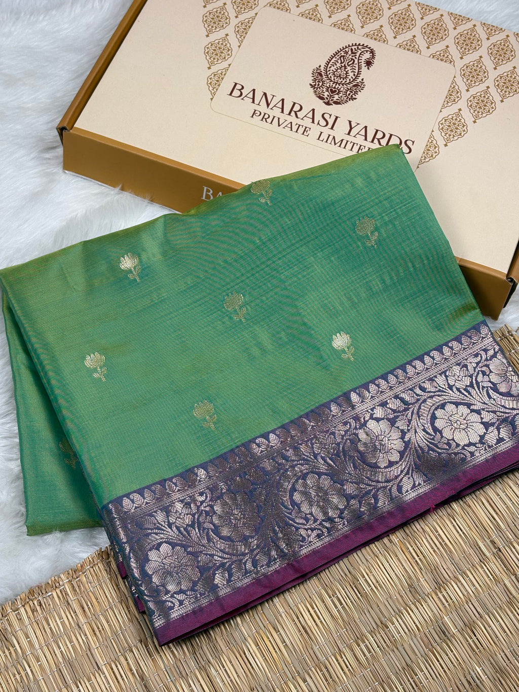 A folded green silk saree with a purple and silver floral border, displayed next to its beige and Banarasi Yards Private Limited box on a white faux fur surface.
