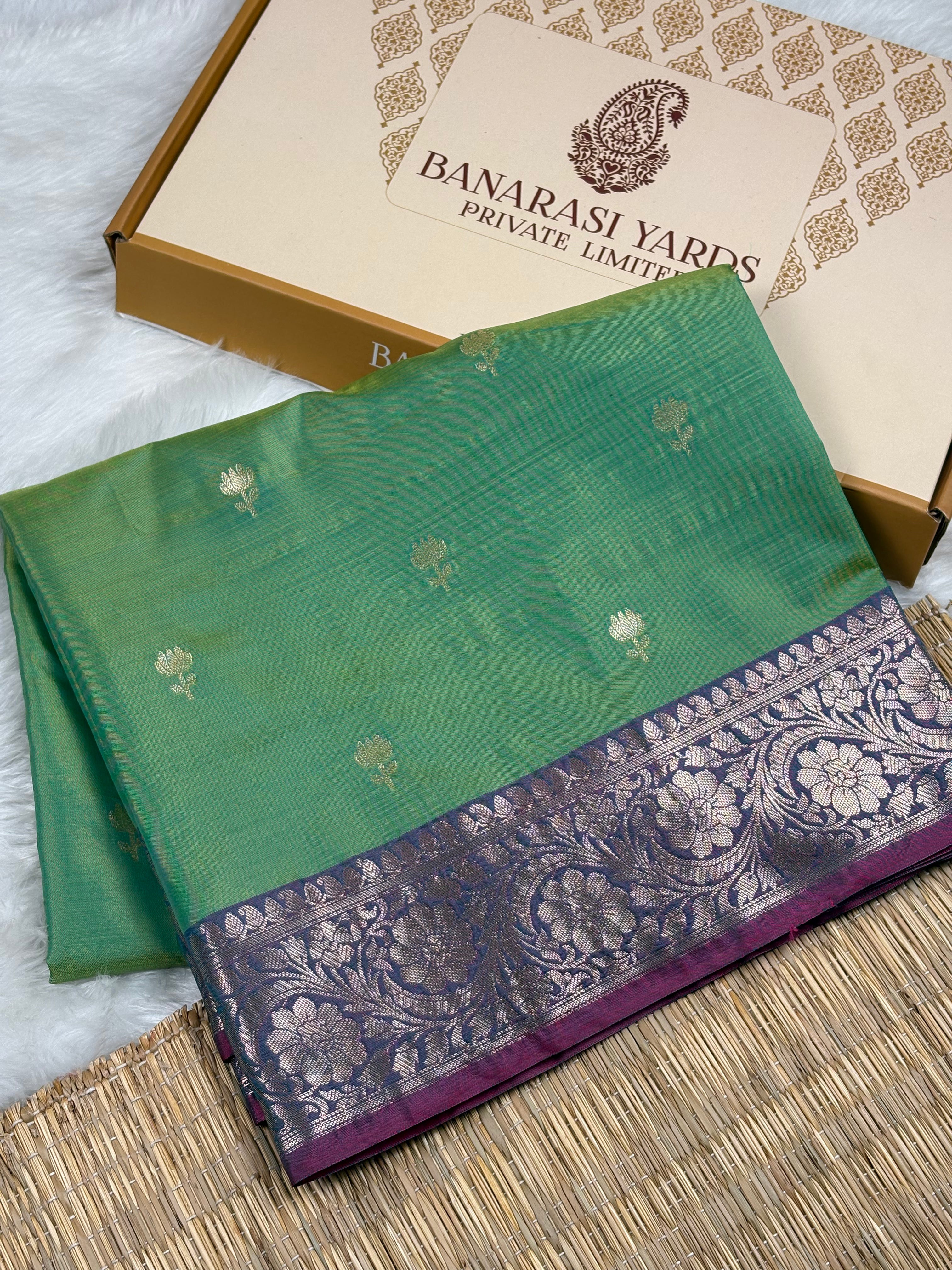 A folded green silk saree with a purple and silver floral border, displayed next to its beige and Banarasi Yards Private Limited box on a white faux fur surface.
