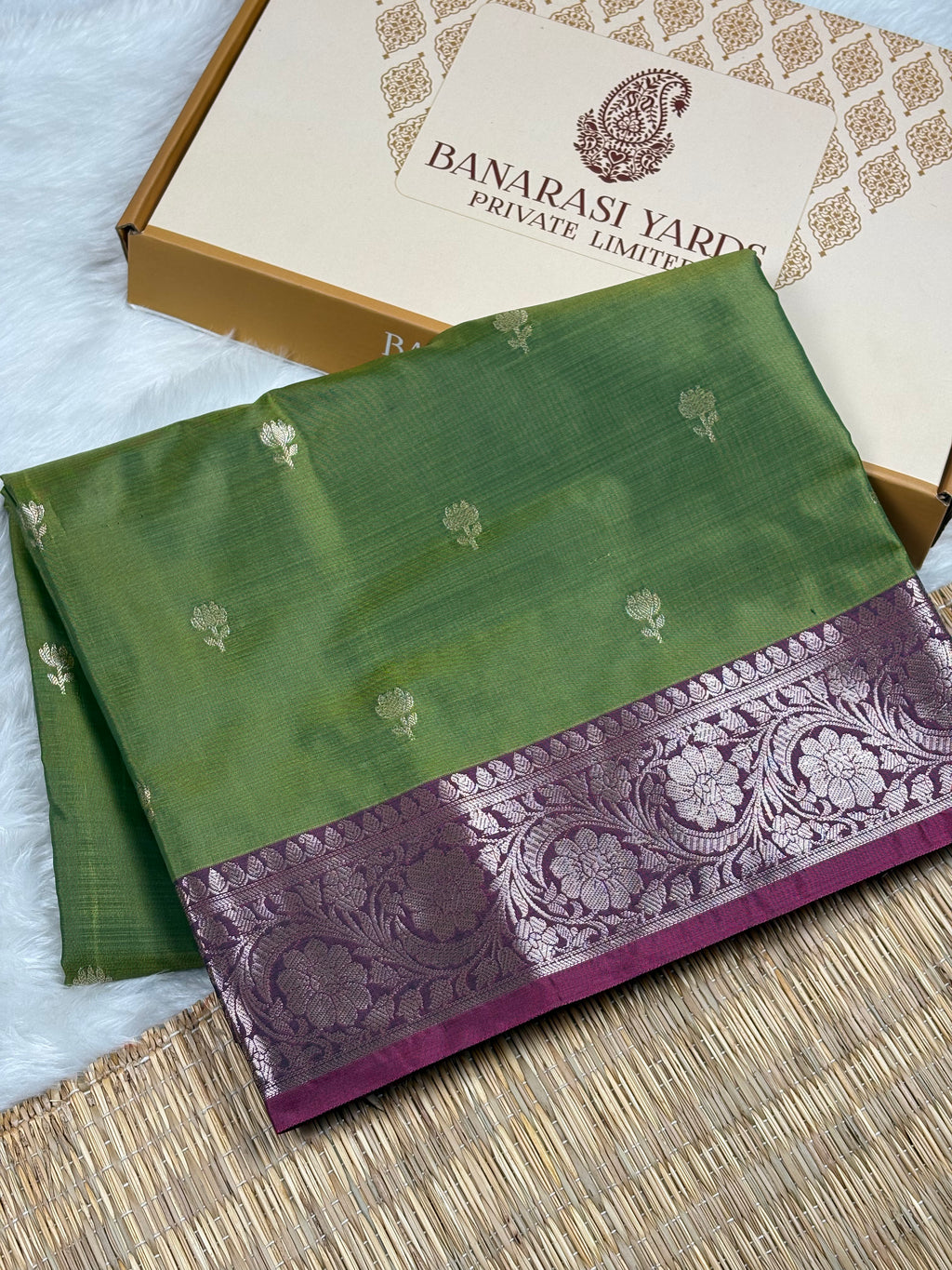 A folded green silk saree with a wide purple border featuring intricate floral zari weaving, displayed next to its beige Banarasi Yards Private Limited box on a woven mat.