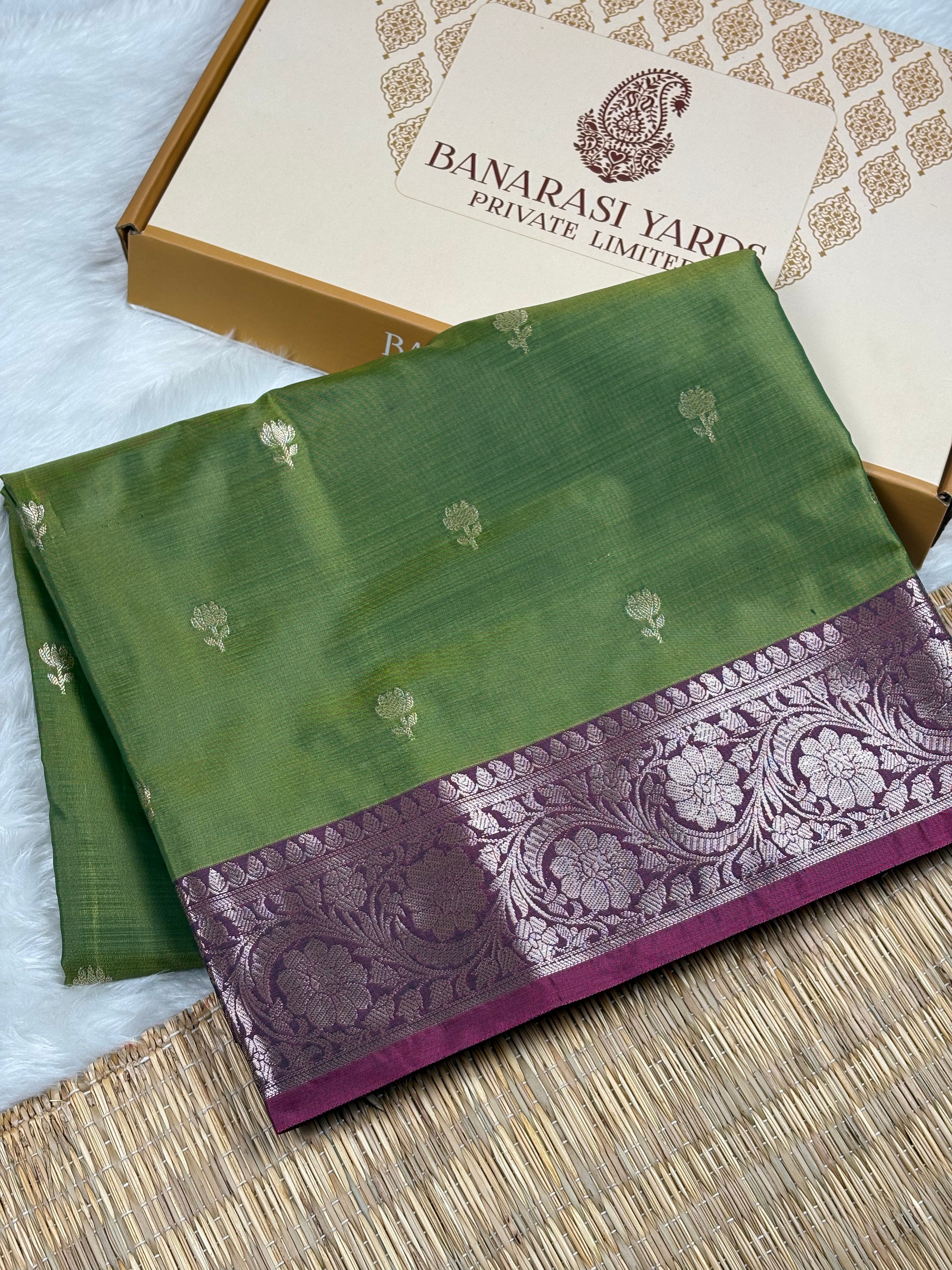 A folded green silk saree with a wide purple border featuring intricate floral zari weaving, displayed next to its beige Banarasi Yards Private Limited box on a woven mat.