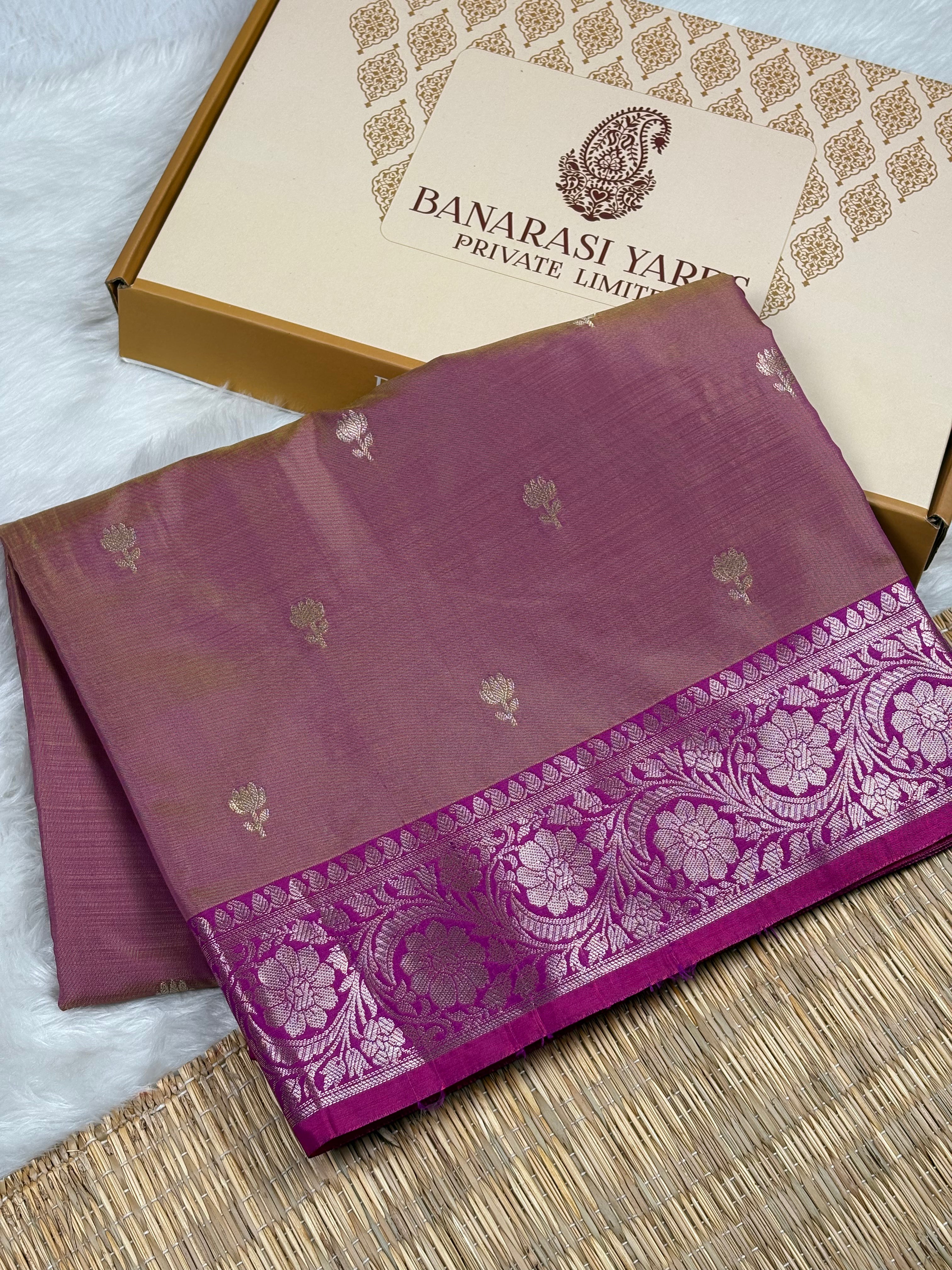 A folded purple silk saree with a floral border, displayed next to its branded packaging box on a woven mat.