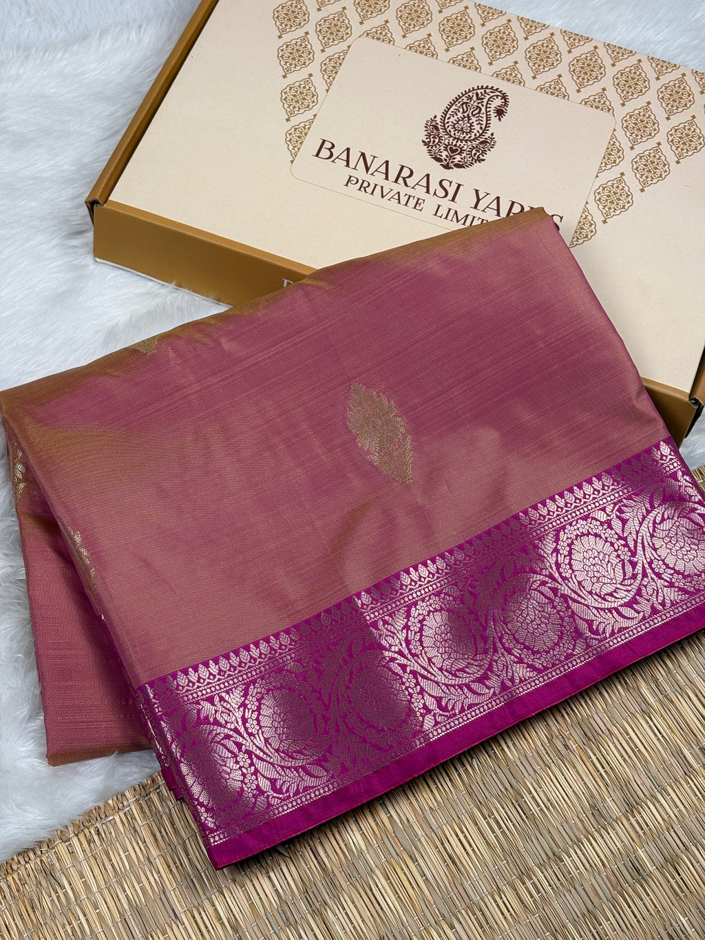 A folded magenta pink silk saree with a wide gold border, displayed next to its beige and gold Banarasi Yards Private Limited box on a woven mat.