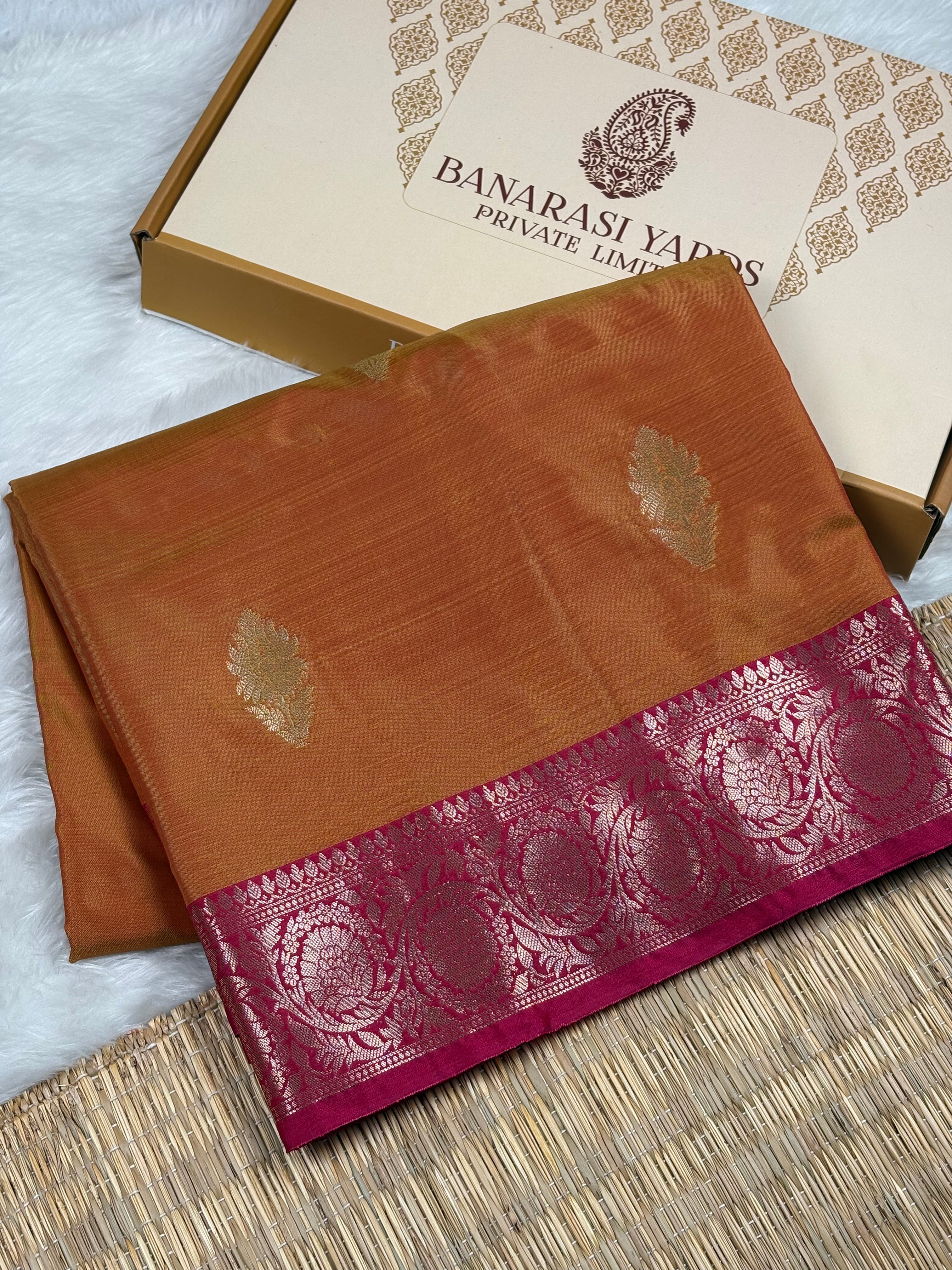 A folded orange-gold saree with a deep magenta border and gold zari motifs, displayed next to its branded Banarasi Yards Private Limited box on a woven mat.