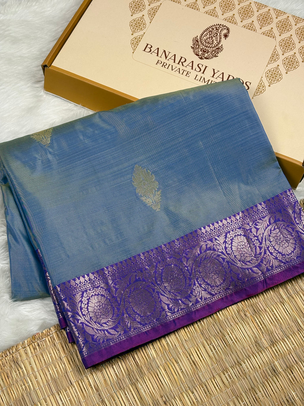 A folded blue saree with a purple border and gold zari motifs, displayed next to its beige Banarasi Yards box on a woven mat.