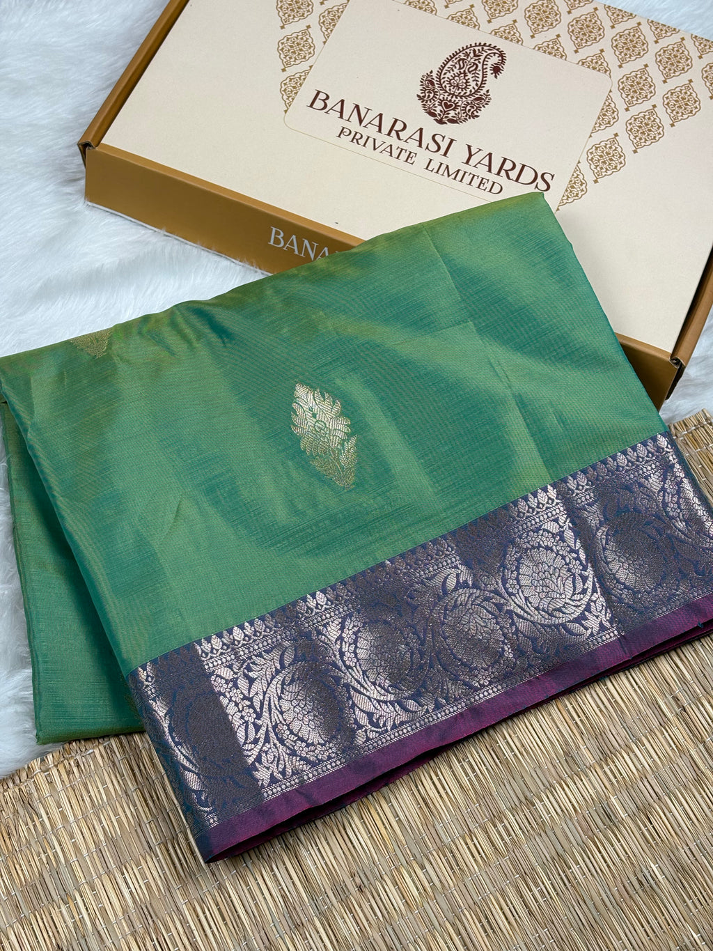 A folded green silk saree with a purple and silver patterned border, displayed next to its beige and gold Banarasi Yards Private Limited box on a woven mat.