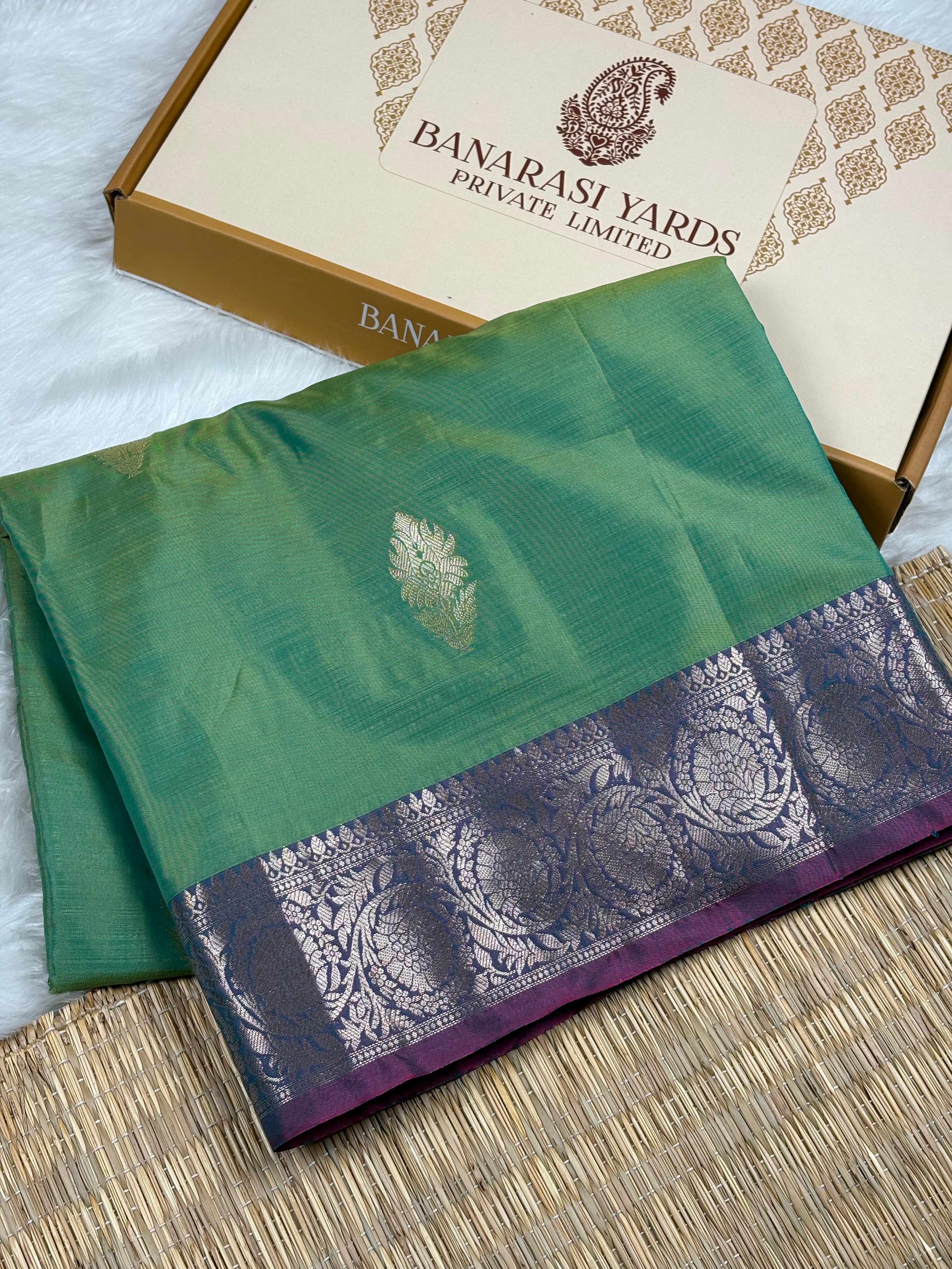 A folded green silk saree with a purple and silver patterned border, displayed next to its beige and gold Banarasi Yards Private Limited box on a woven mat.