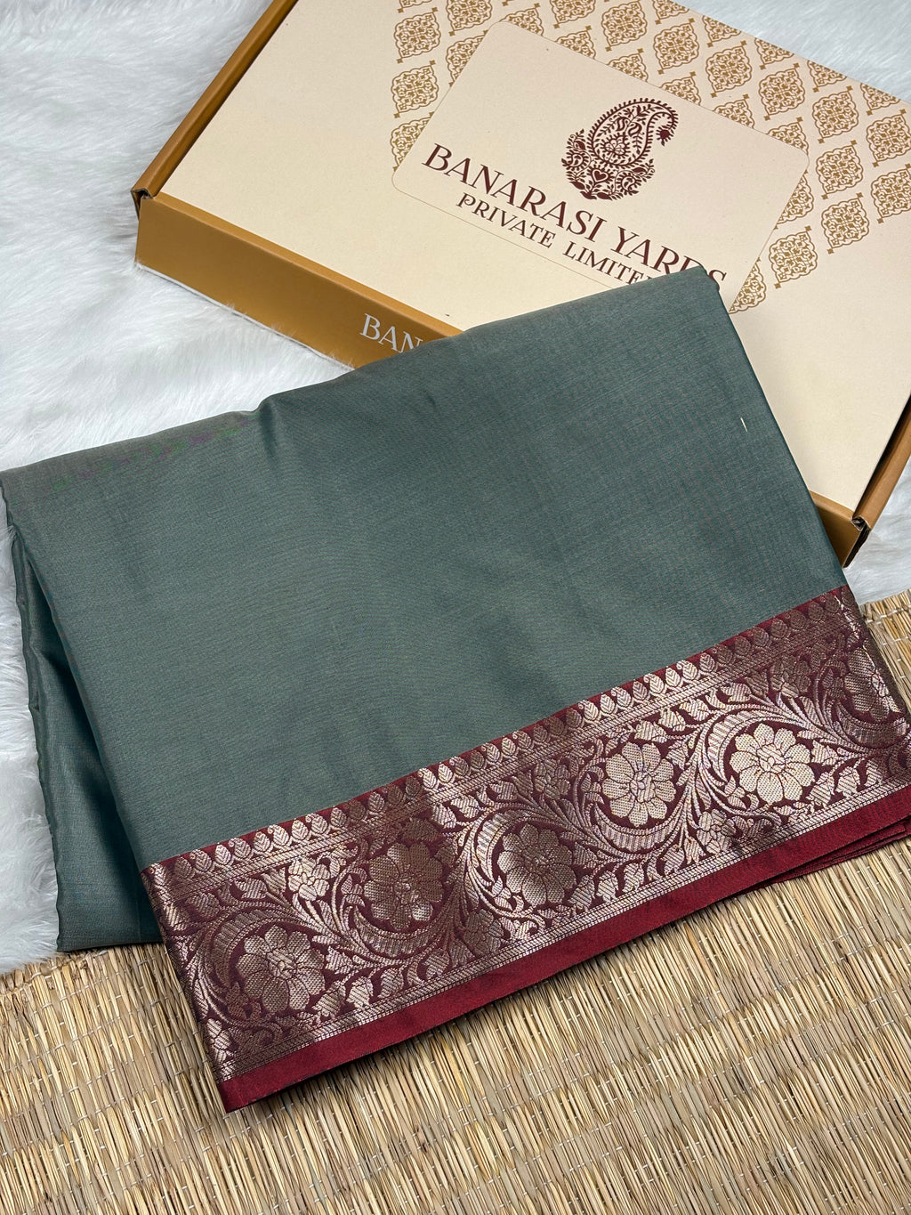 A folded dark gray-green saree with a wide gold floral border, displayed next to its Banarasi Yards Private Limited box on a woven mat.
