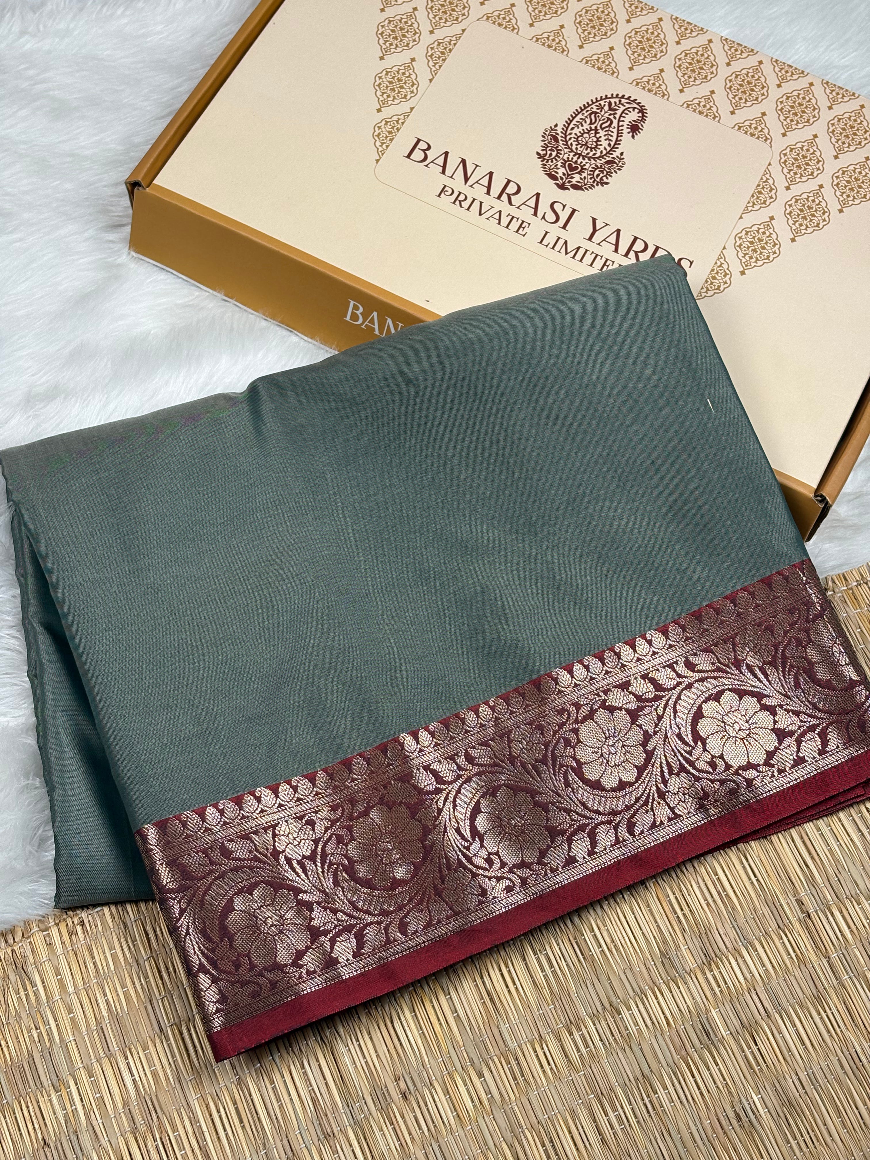 A folded dark gray-green saree with a wide gold floral border, displayed next to its Banarasi Yards Private Limited box on a woven mat.