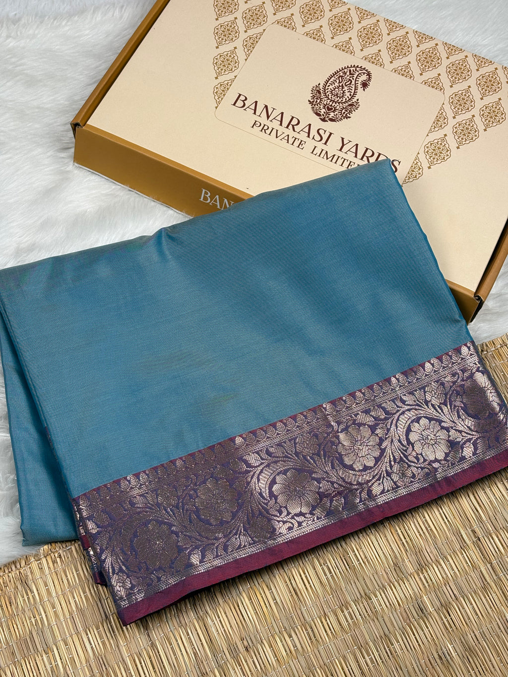 A folded blue saree with a wide purple and gold floral border, displayed next to its beige and gold Banarasi Yards box on a woven mat.