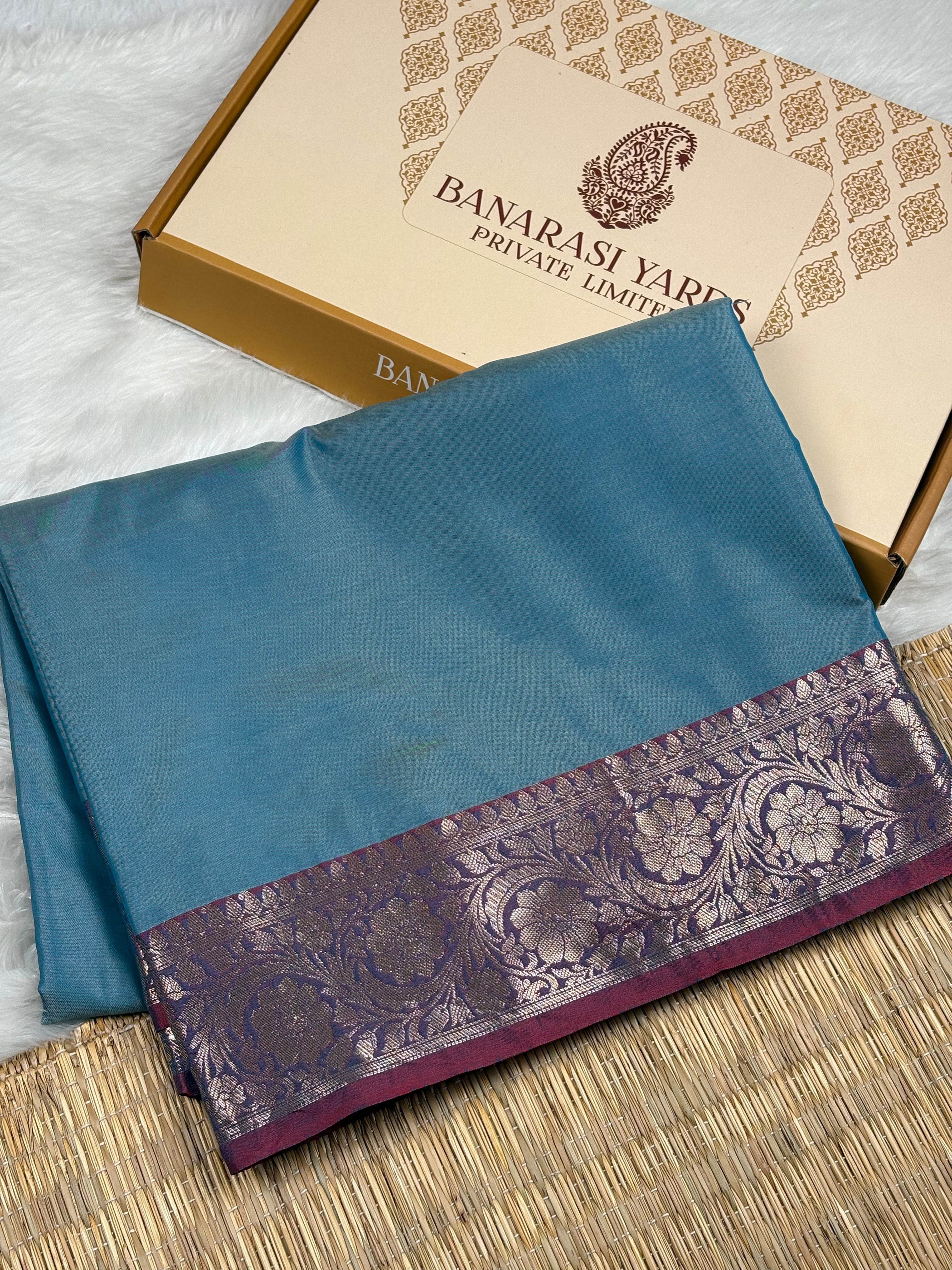 A folded blue saree with a wide purple and gold floral border, displayed next to its beige and gold Banarasi Yards box on a woven mat.