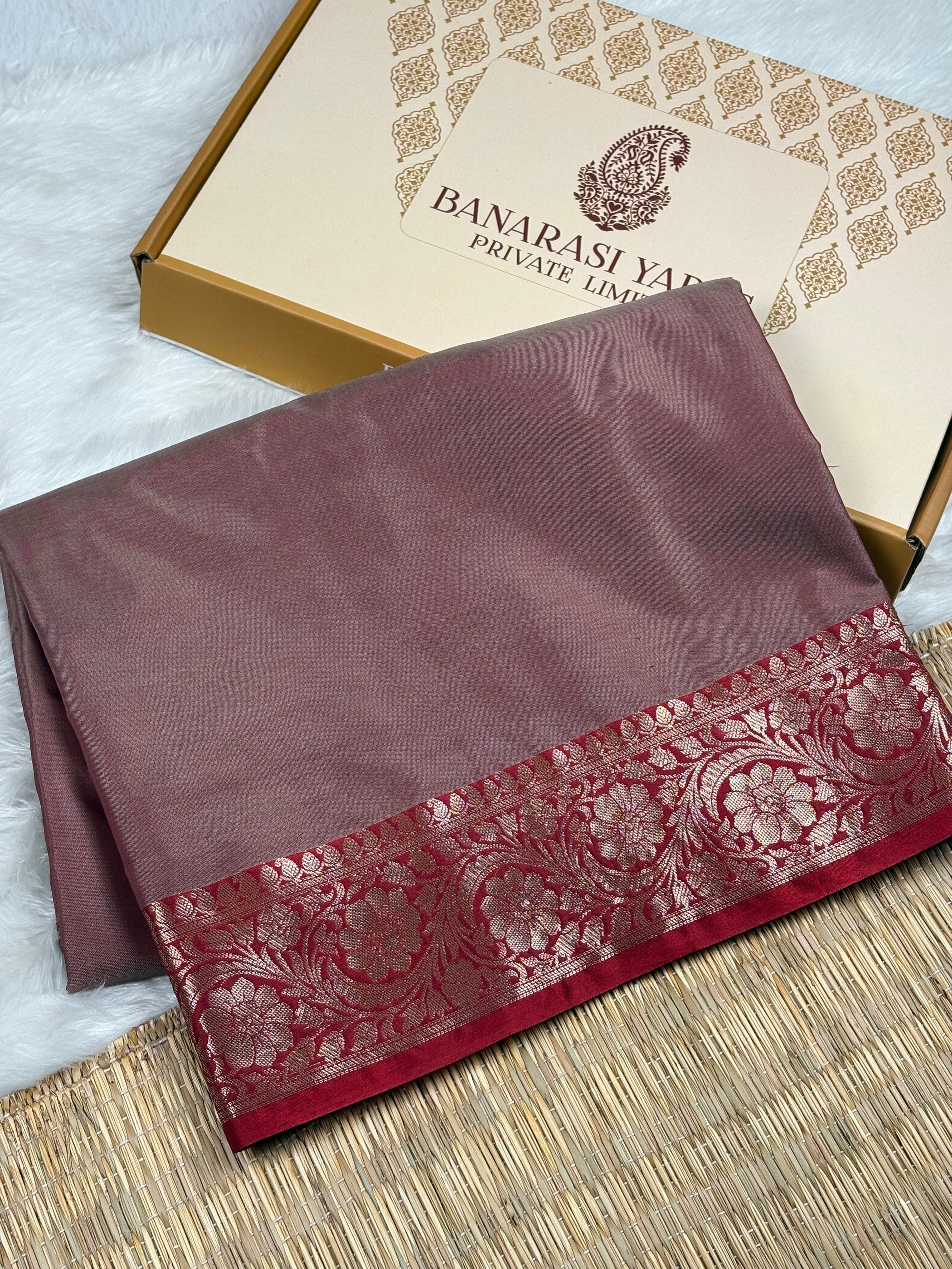 A folded dusty rose silk saree with a red and gold floral zari border, displayed next to its branded Banarasi Yards Private Limited gift box on a woven mat.