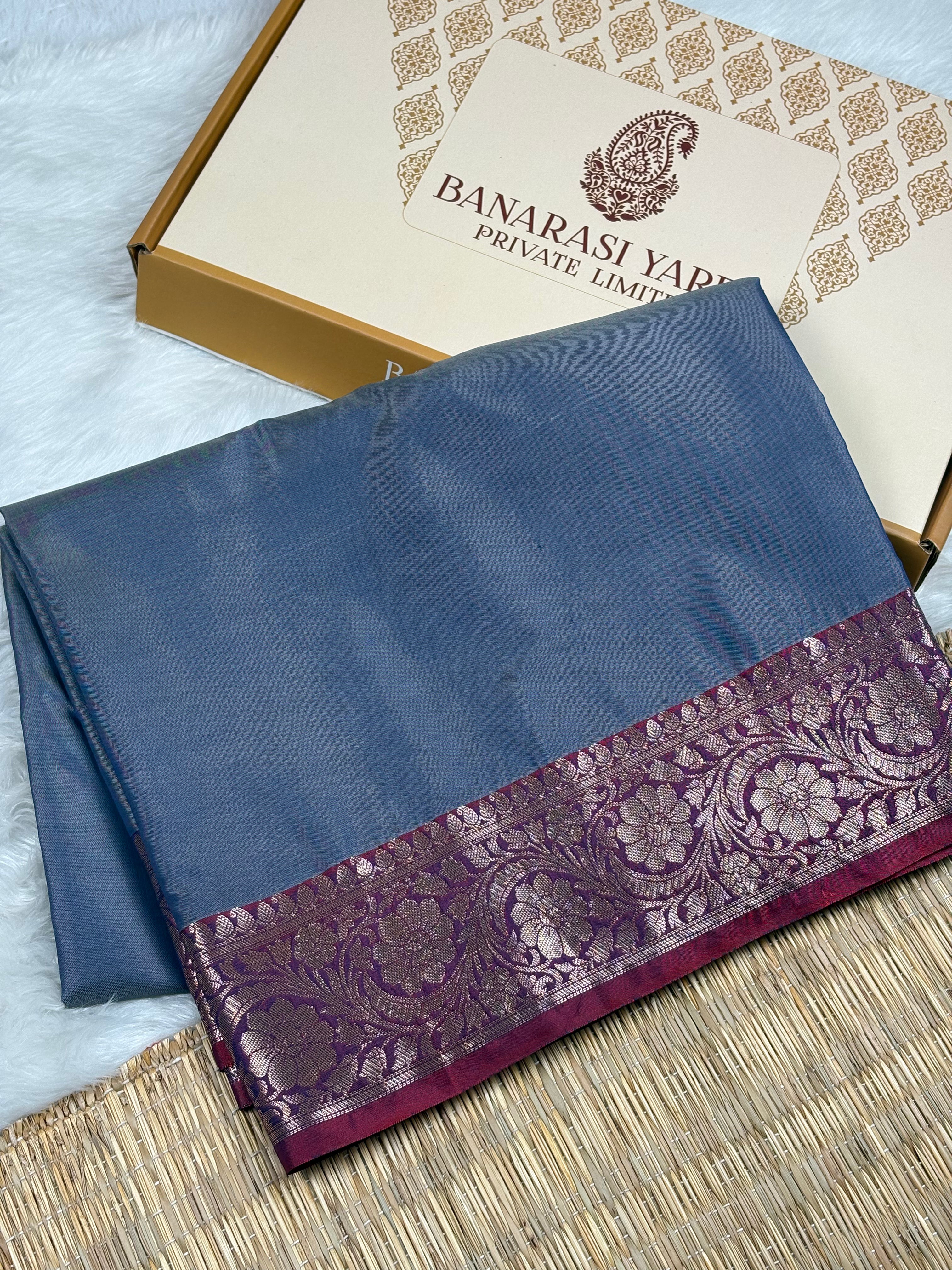 A folded blue saree with a wide purple and zari border, displayed next to its branded packaging box on a textured mat.