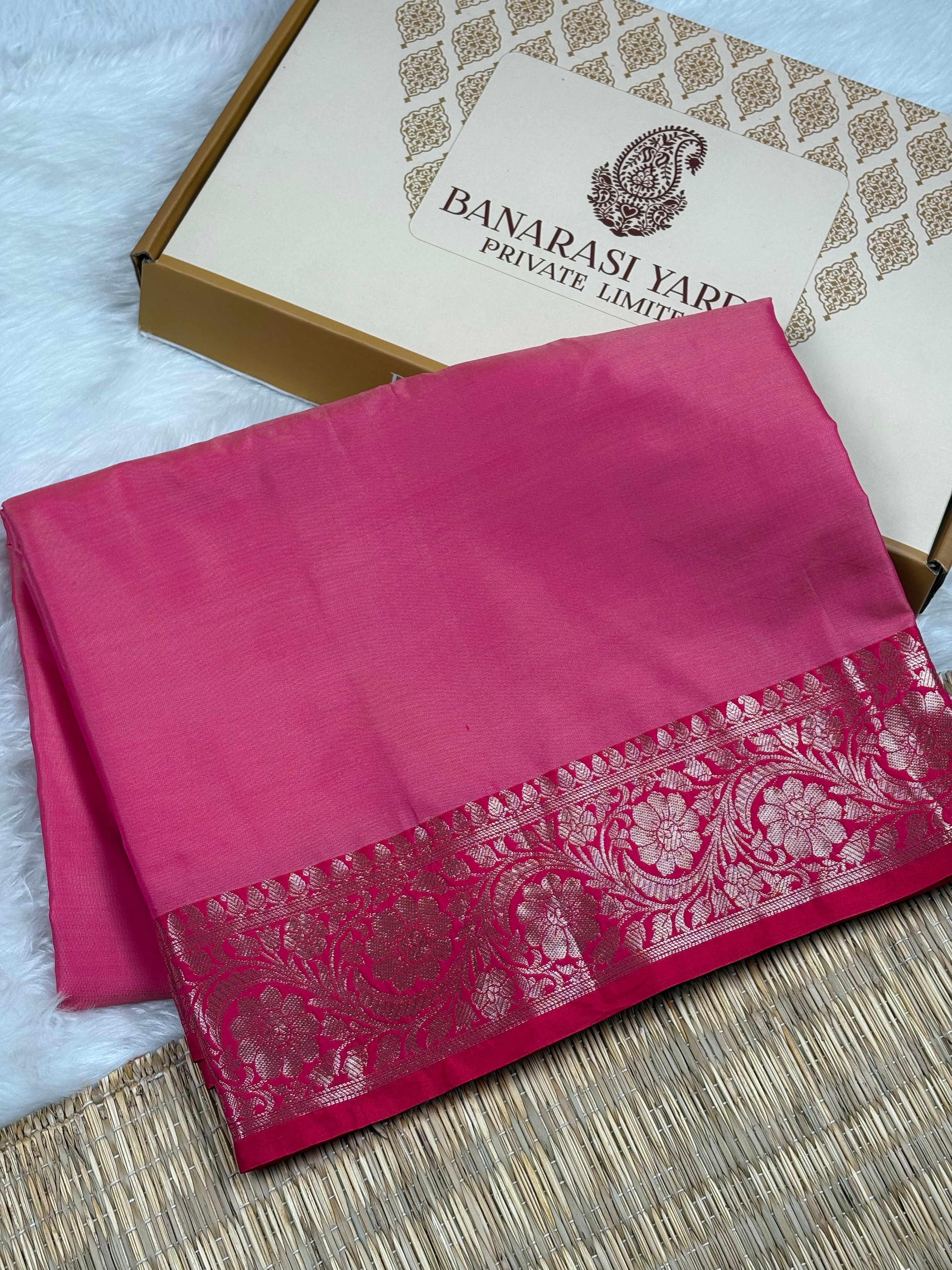 A folded magenta pink Banarasi silk saree with a gold floral border weaving, displayed next to its 'Banarasi Yards Private Limited' box on a woven mat.