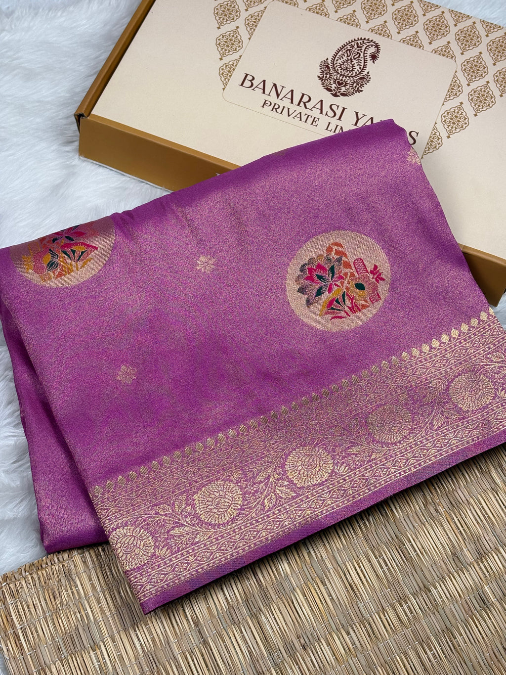 A folded purple soft tishu silk saree with a gold zari border and floral motifs, displayed next to its branded gift box on a woven mat.
