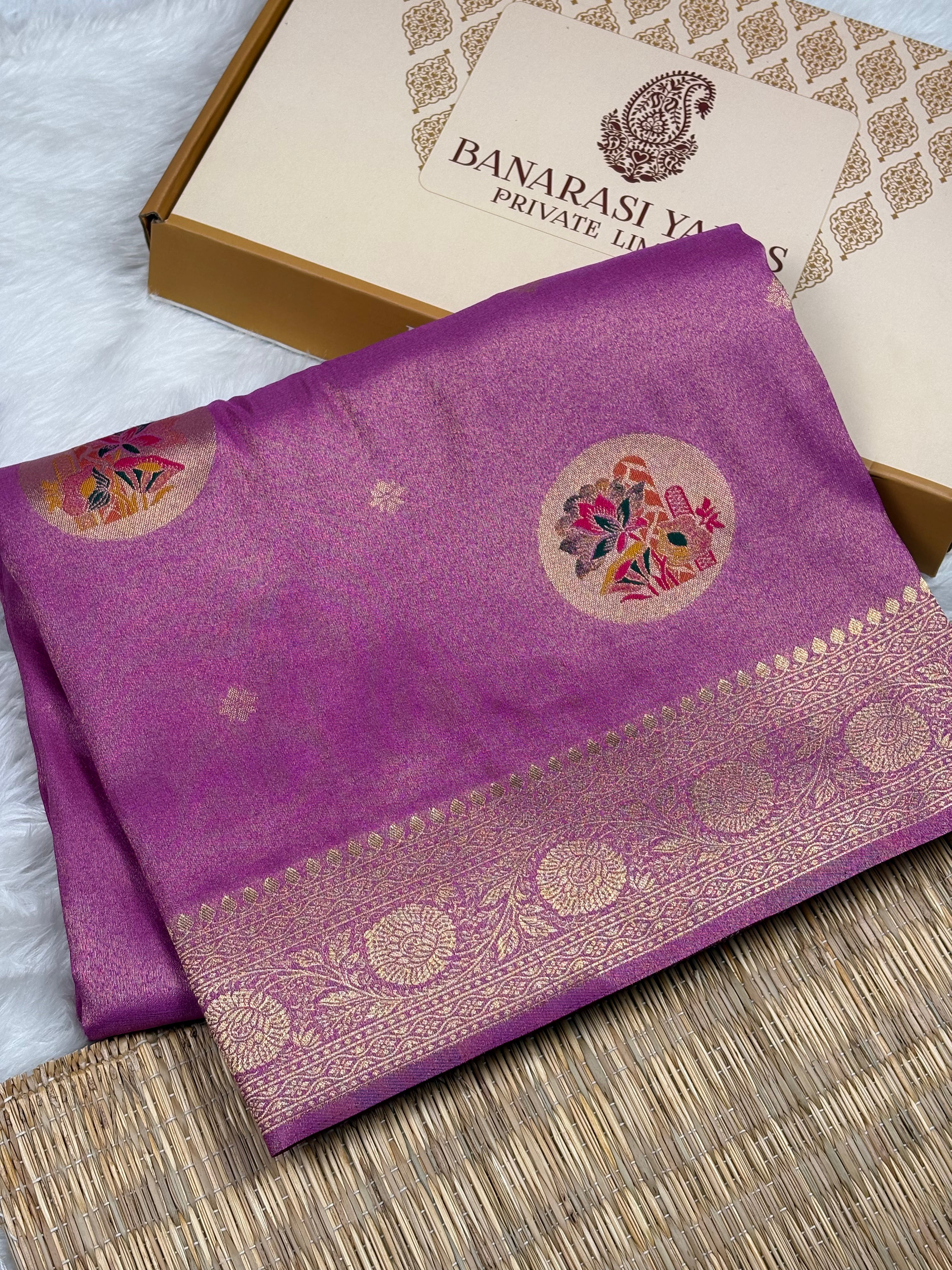 A folded purple soft tishu silk saree with a gold zari border and floral motifs, displayed next to its branded gift box on a woven mat.