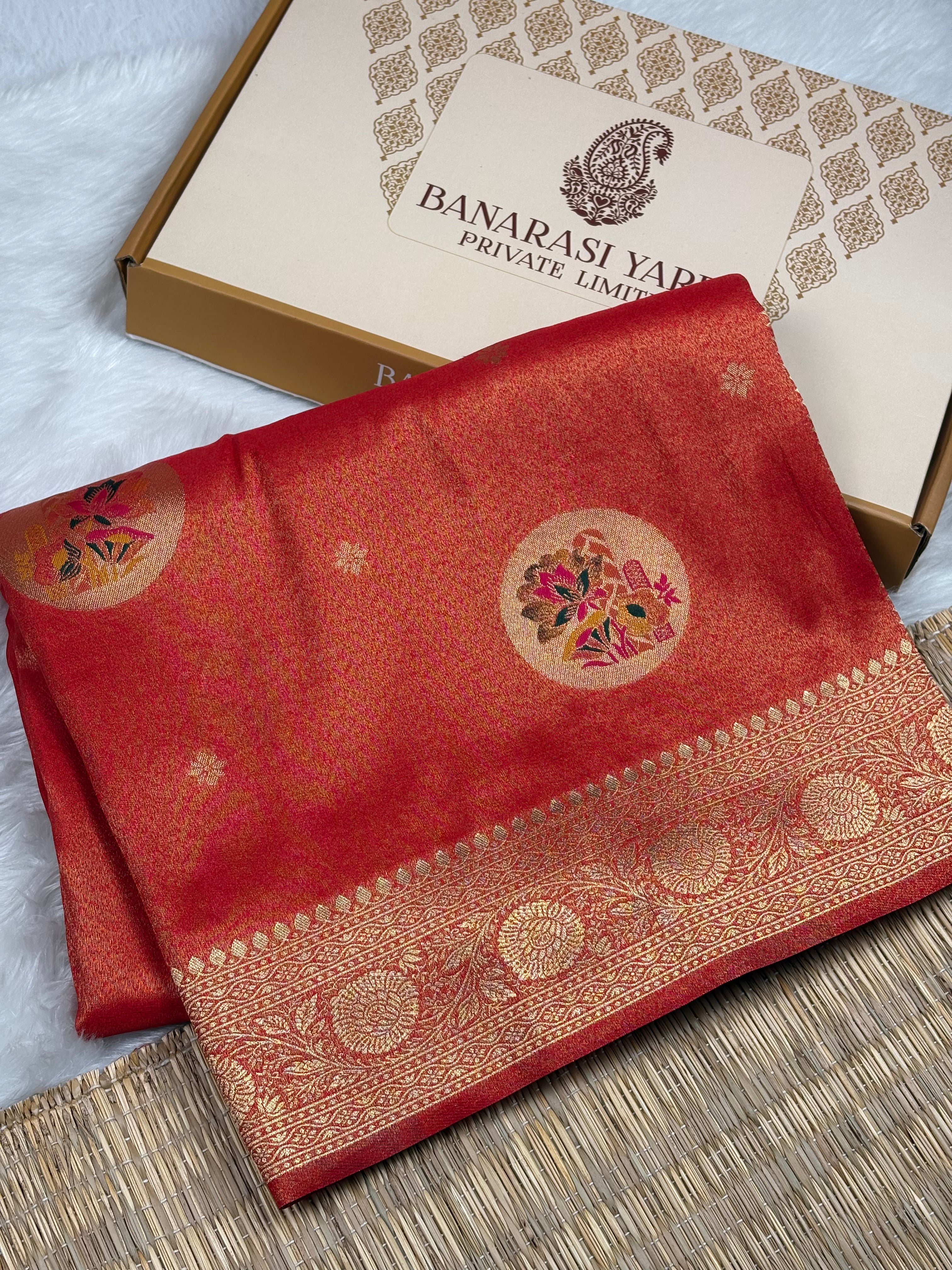 A folded red soft tishu Banarasi silk saree with intricate gold zari weaving and floral motifs, displayed next to its branded packaging box on a woven mat.