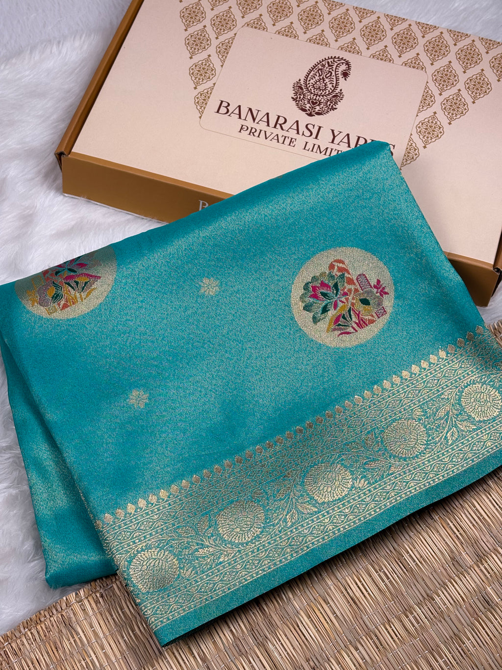 A folded turquoise blue soft tishu silk saree with intricate gold zari work and floral motifs, displayed next to its beige and gold Banarasi Yards Private Limited gift box on a white faux fur surface.