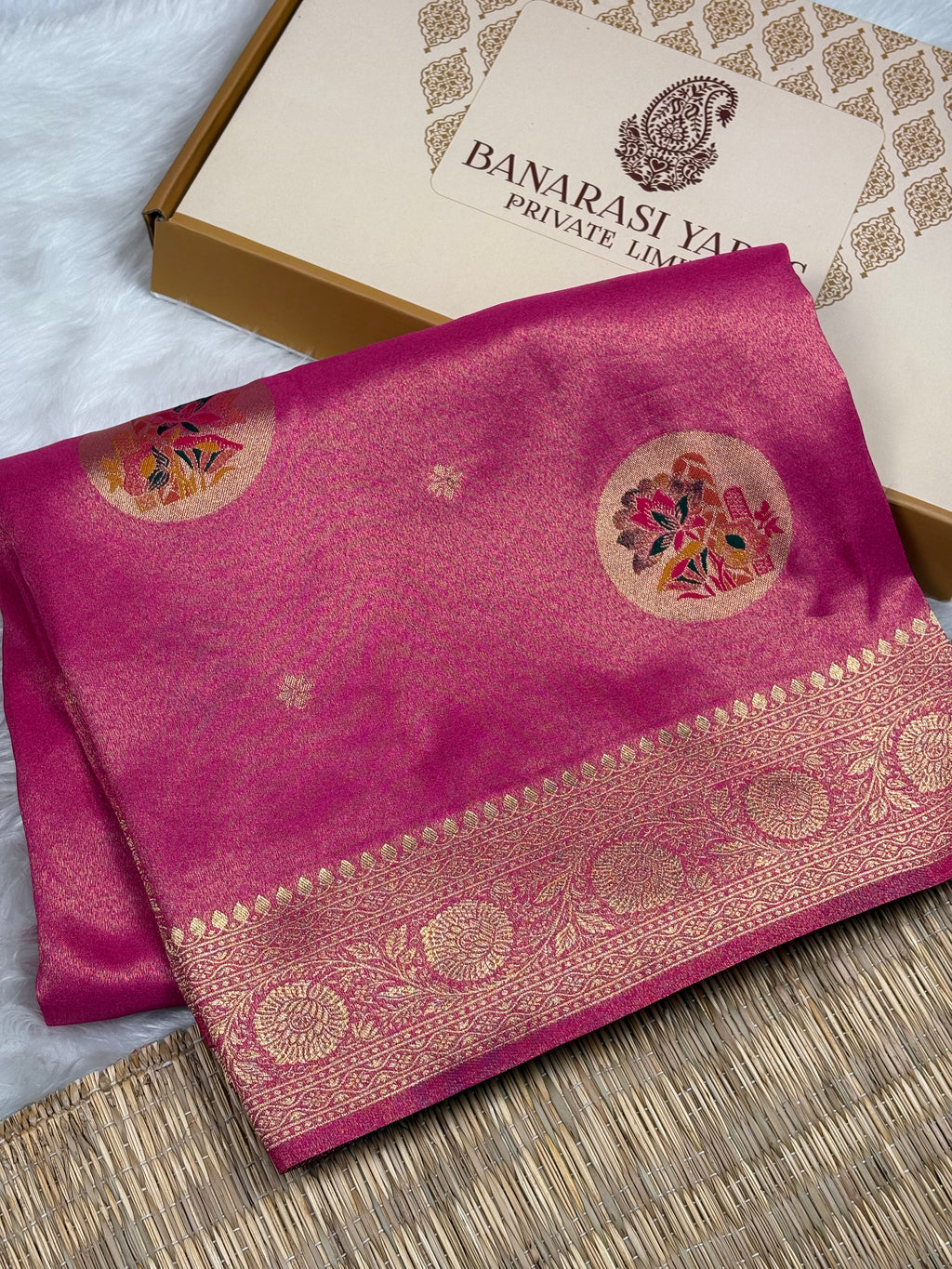 A folded magenta pink soft tishu silk saree with a gold zari border and floral motifs, displayed next to its branded packaging box on a woven mat.