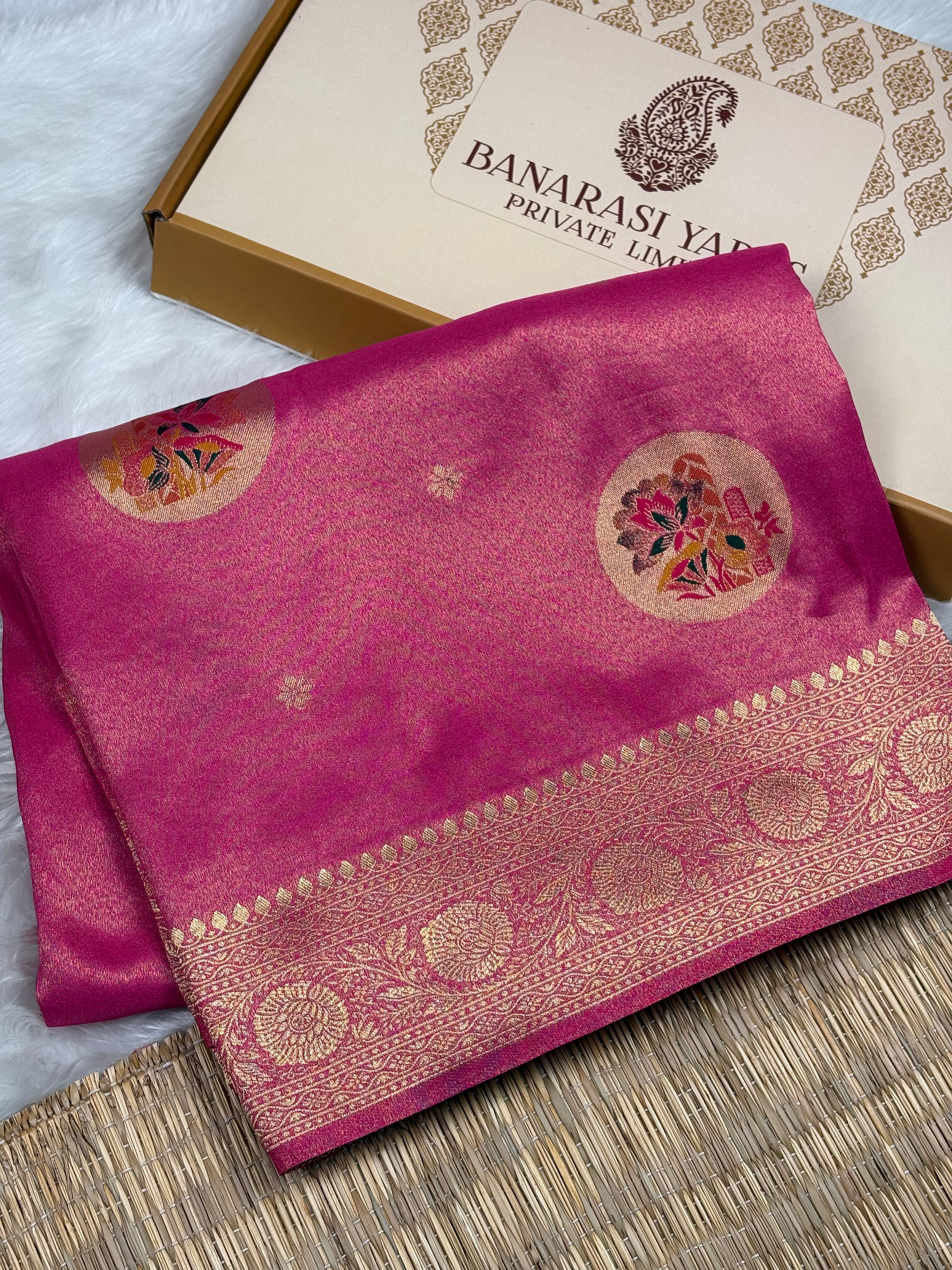 A folded magenta pink soft tishu silk saree with a gold zari border and floral motifs, displayed next to its branded packaging box on a woven mat.