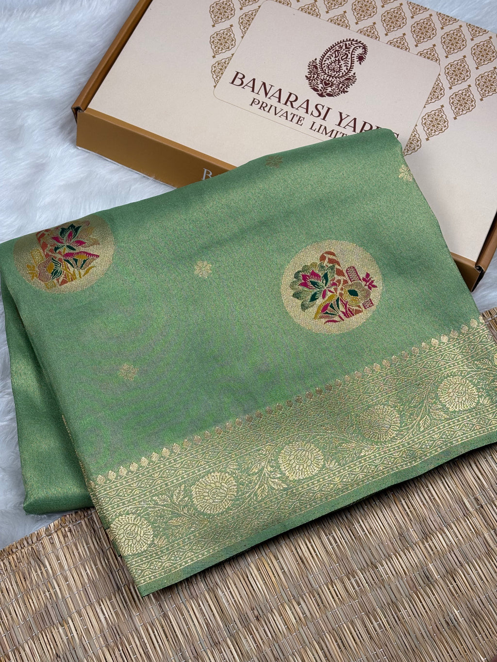 A folded sage green soft tishu Banarasi silk saree with gold zari weaving and floral motifs, displayed next to its packaging box on a woven mat.
