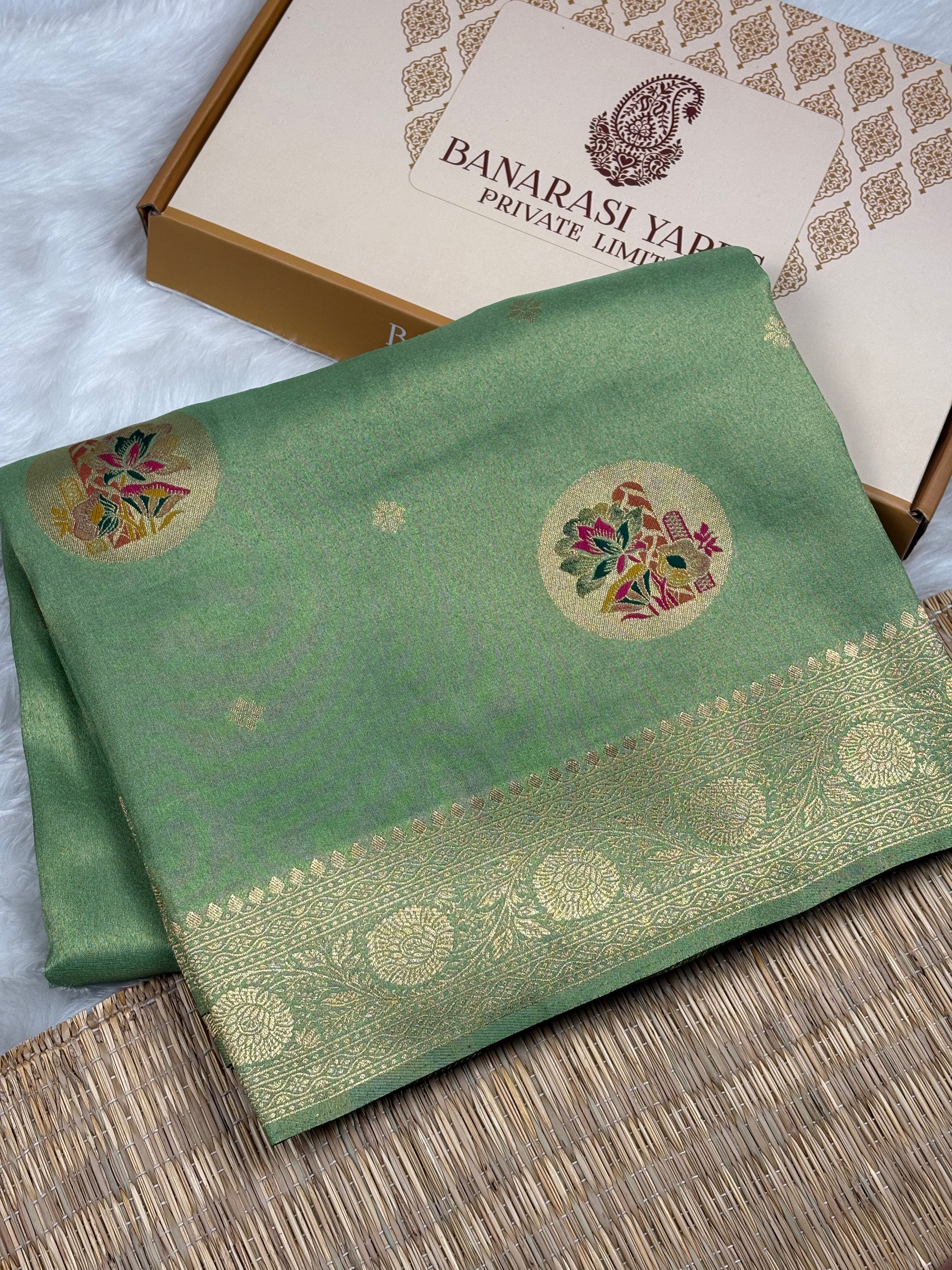 A folded sage green soft tishu Banarasi silk saree with gold zari weaving and floral motifs, displayed next to its packaging box on a woven mat.
