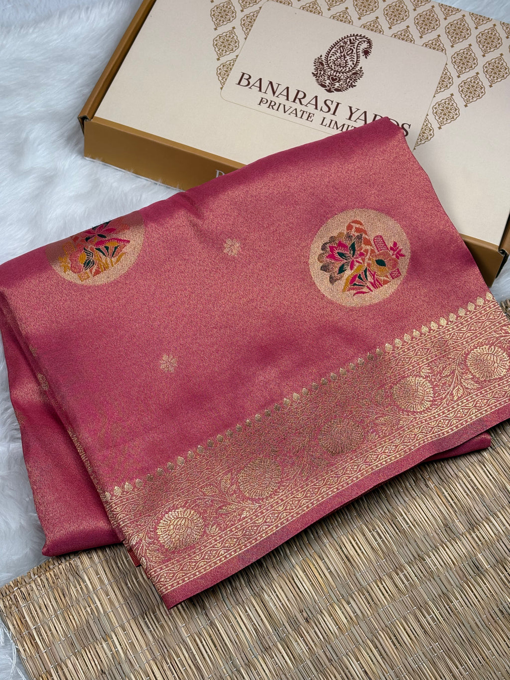 A folded pink Banarasi soft tishu silk saree with gold zari weaving and floral motifs, displayed next to its branded packaging box on a textured mat.