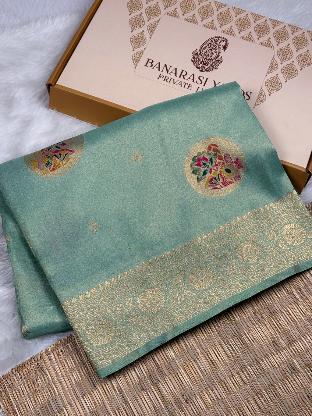 A folded sage green soft tishu silk saree with gold zari borders and floral motifs, displayed next to its beige Banarasi yards Private Limited box on a white faux fur surface and woven mat.