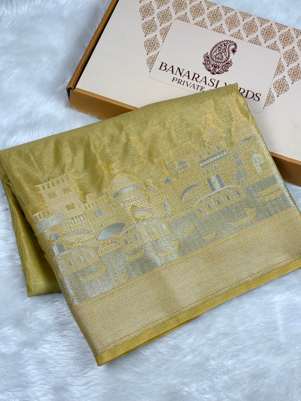 A folded mustard yellow silk saree with alfi mina cityscape pattern and a wide border, displayed next to its branded Banarasi yards Private limited box on a white faux fur surface.