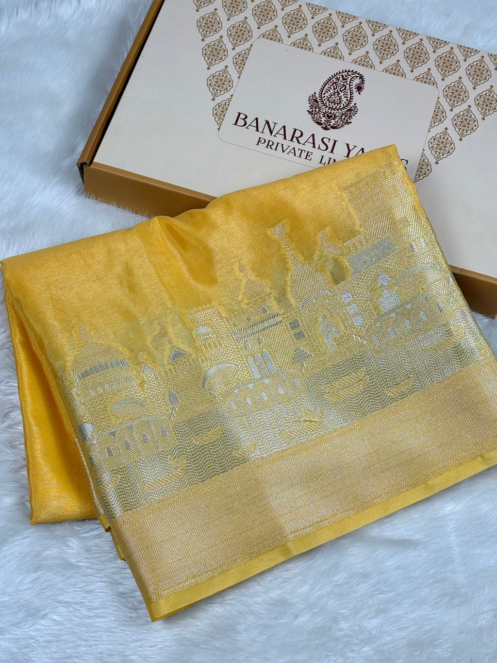 A folded yellow Banarasi silk saree with alfi mina cityscape pattern and a wide border, displayed next to its branded packaging box on a white textured background.
