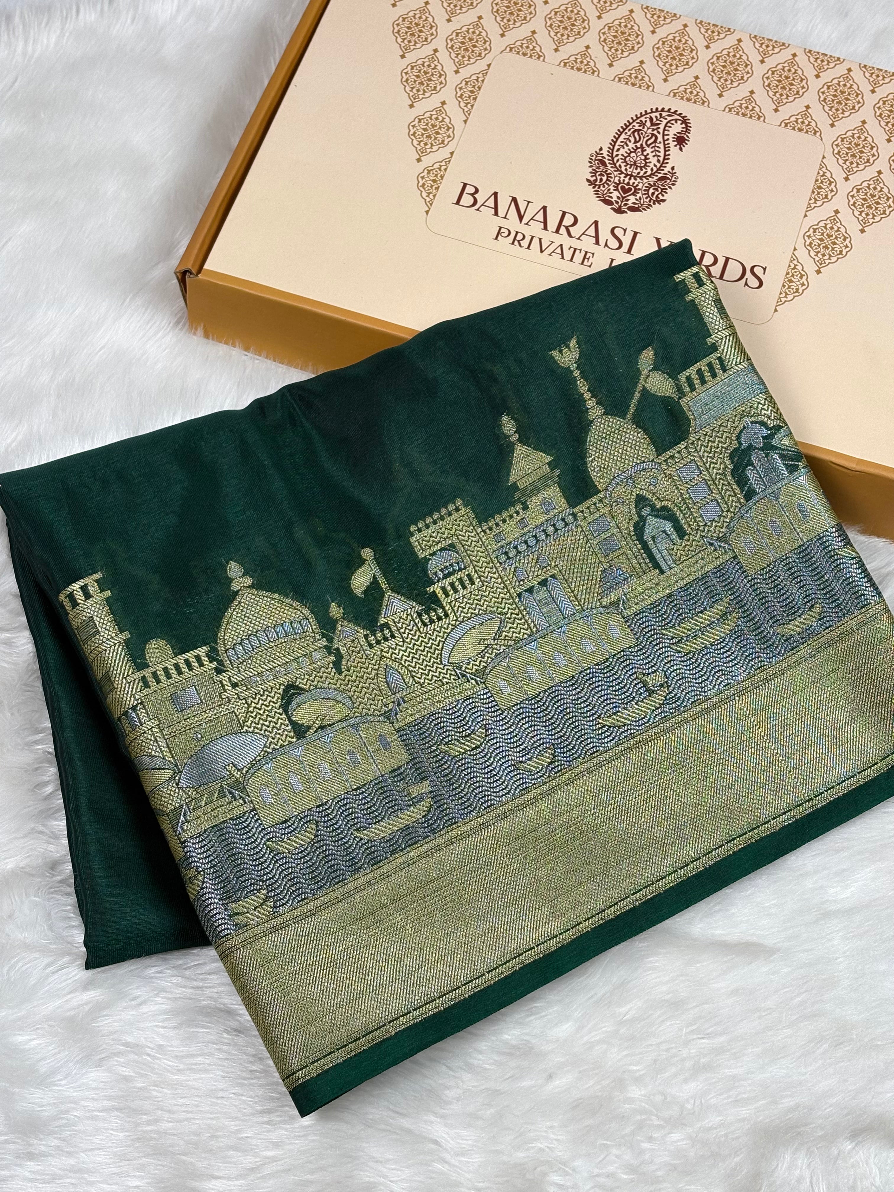 A folded dark green silk saree with intricate alfi mina zari weaving depicting a palace scene, displayed next to its Banarasi Yards Private Limited gift box on a white faux fur background.