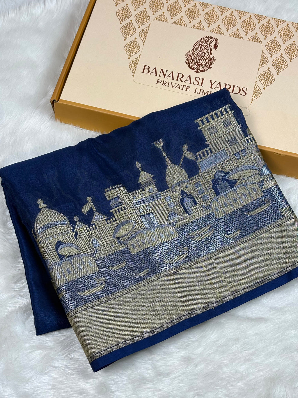 A folded dark blue Banarasi silk saree with a detailed alfi mina zari architectural border weaving, displayed next to its Banarasi Yards gift box on a white faux fur background.