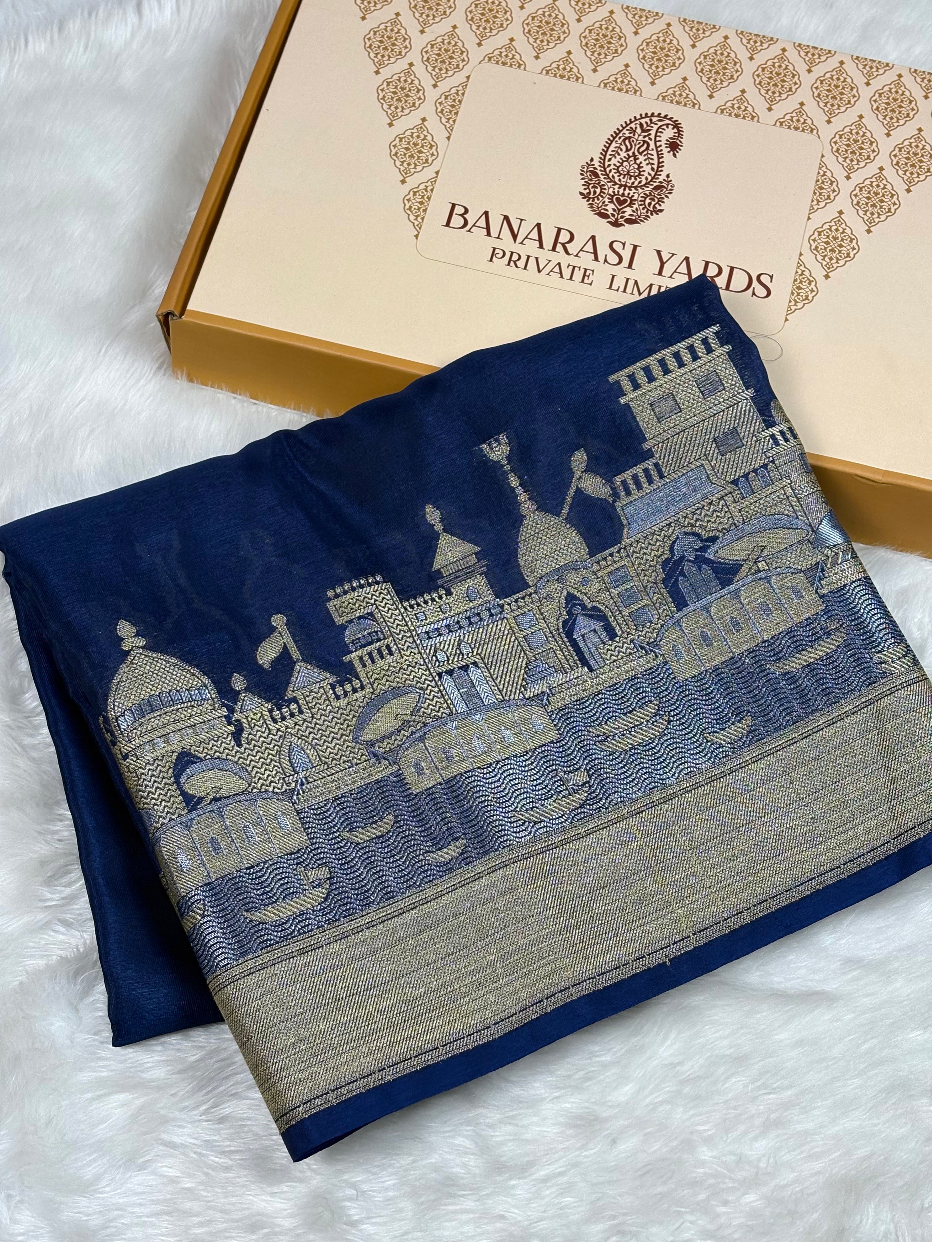 A folded dark blue Banarasi silk saree with a detailed alfi mina zari architectural border weaving, displayed next to its Banarasi Yards gift box on a white faux fur background.