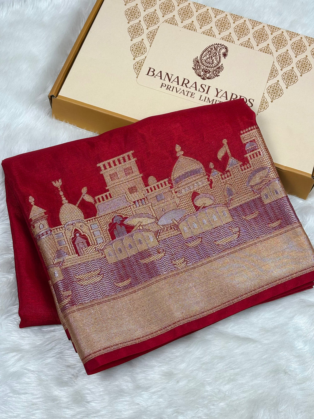 A folded red Banarasi silk saree with a detailed alfi mina zari architectural border, displayed next to its Banarasi Yards Private Limited gift box on a white faux fur background.